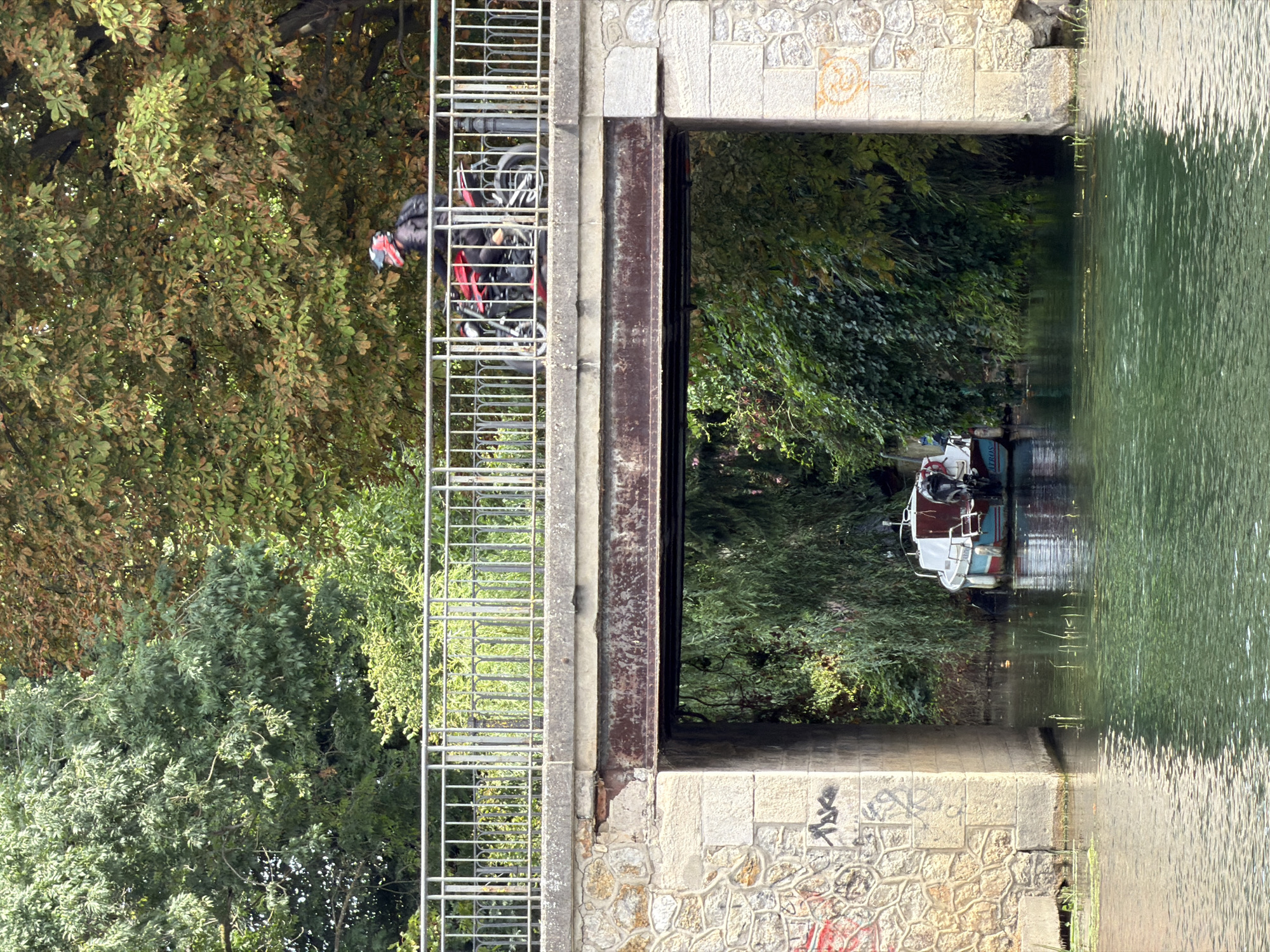 The image depicts a bridge crossing a canal in Joinville-le-Pont, France. A person wearing a red and black helmet rides a red motorcycle across the bridge, with a black backpack visible. The bridge itself is constructed of stone and has a metal railing. Beneath the bridge, a small white boat with red and blue trim is moored in the canal. The canal banks are covered in dense green foliage, and trees with autumnal leaves are visible on either side of the canal. The water in the canal is a murky green, reflecting the surrounding vegetation. Graffiti is present on the stone pillars of the bridge.