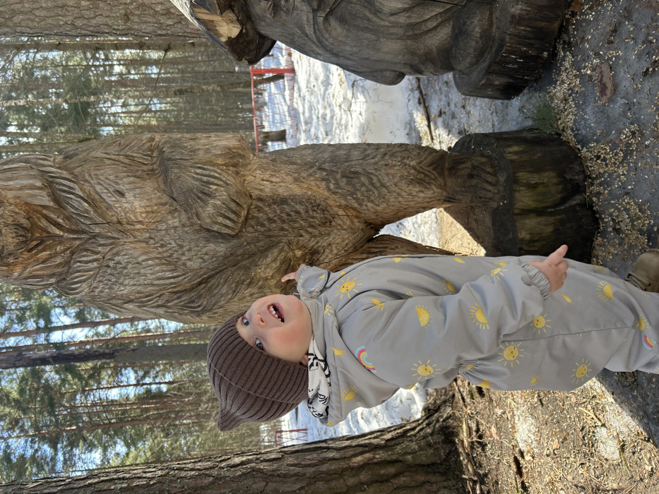 A young child, wearing a brown knit beanie and a grey full-body jumpsuit with yellow sun and rainbow patterns, stands in a wooded area in Dubna, Russia. The child's right index finger points towards the chest area of a large, carved wooden sculpture depicting a standing bear with textured fur. The child's head is tilted back, mouth open, looking upwards towards the sculpture. A second, partially visible carved wooden figure is present to the right. The ground exhibits patches of melting snow, bare earth, and scattered wood shavings around the sculpture bases. Tall, slender coniferous trees form a dense background, through which a red metal structure, possibly a playground element, is discernible.