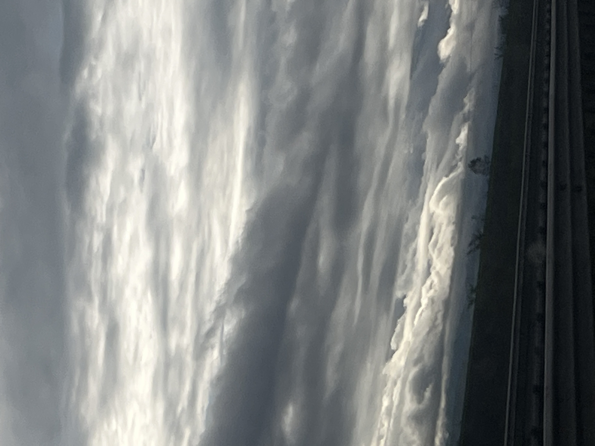 The image, presented with a vertical orientation, captures a sky filled with layered clouds and a dark, prominent structure on the right. If viewed horizontally, the scene reveals an expansive sky dominated by streaky, elongated clouds varying in shades from bright white to dark gray. The clouds exhibit a textured, rippled, or wave-like pattern, suggesting a dynamic weather system, possibly altostratus or cirrocumulus formations.
Bright areas within the clouds indicate the presence of sunlight, making it evident that the photograph was taken during the daytime, likely under overcast conditions.
On the right side of the frame, a significant portion is occupied by a tall, dark, vertical mass. This appears to be an artificial structure, possibly a large building or a series of interconnected structures, distinguishable by its straight vertical lines and dark silhouette. Faint lighter details are discernible against this dark mass, which could be distant vegetation or architectural elements. The transition between the luminous clouds and the dark structure is sharp and distinct.
No people or specific activities are visible in the image. The weather is cloudy. No visible text is present. The precise location or specific time of day beyond "daytime" cannot be determined from the visual cues.