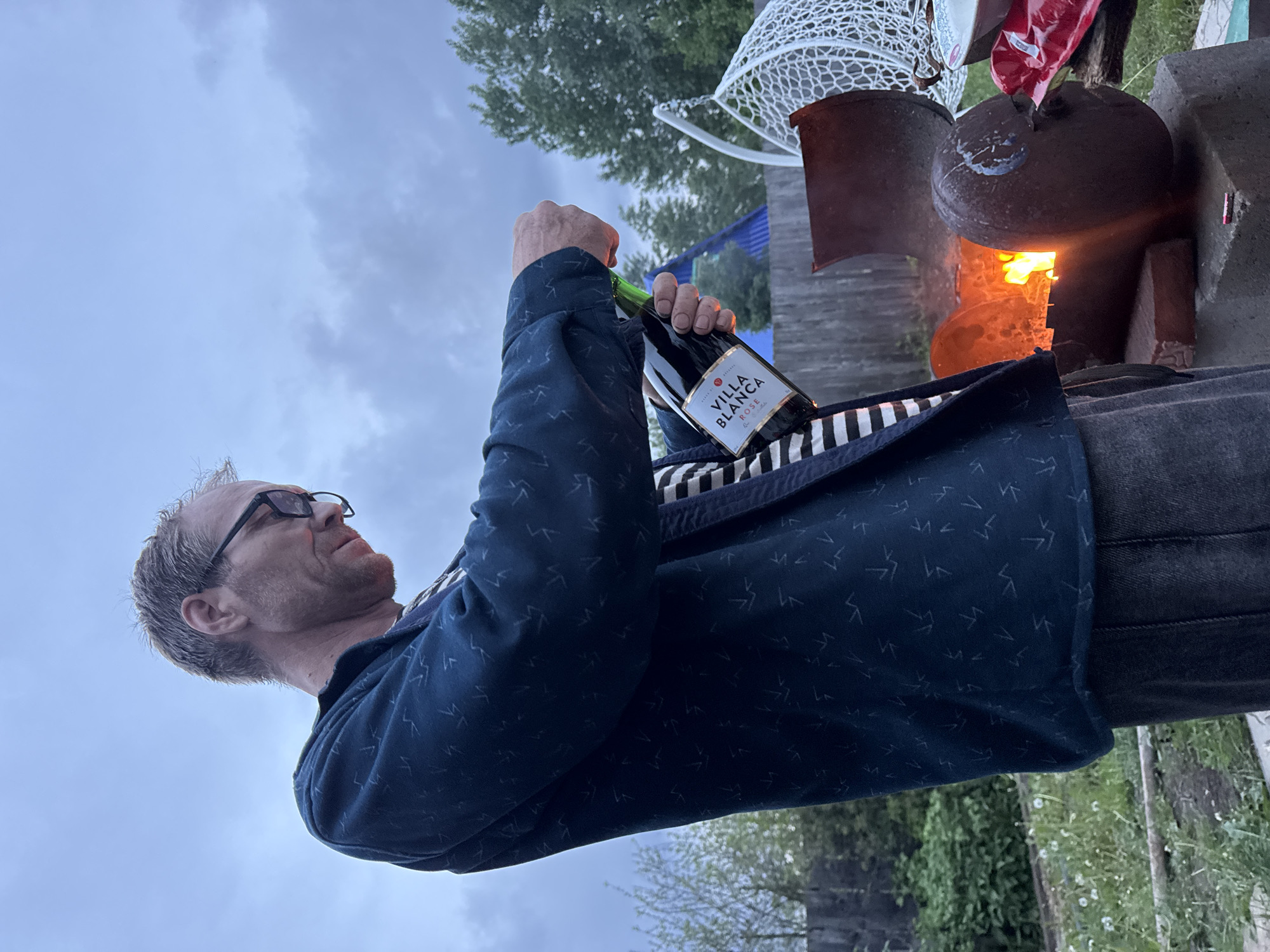 A man with eyeglasses is opening a bottle of Villa Blanca rosé. The bottle is held over a fire pit constructed from bricks and metal objects, and a grill with a white wire frame is present near the pit. The location appears to be outdoors with trees and cloudy skies visible in the background. The location is the city of Chaikovsky, Russia.