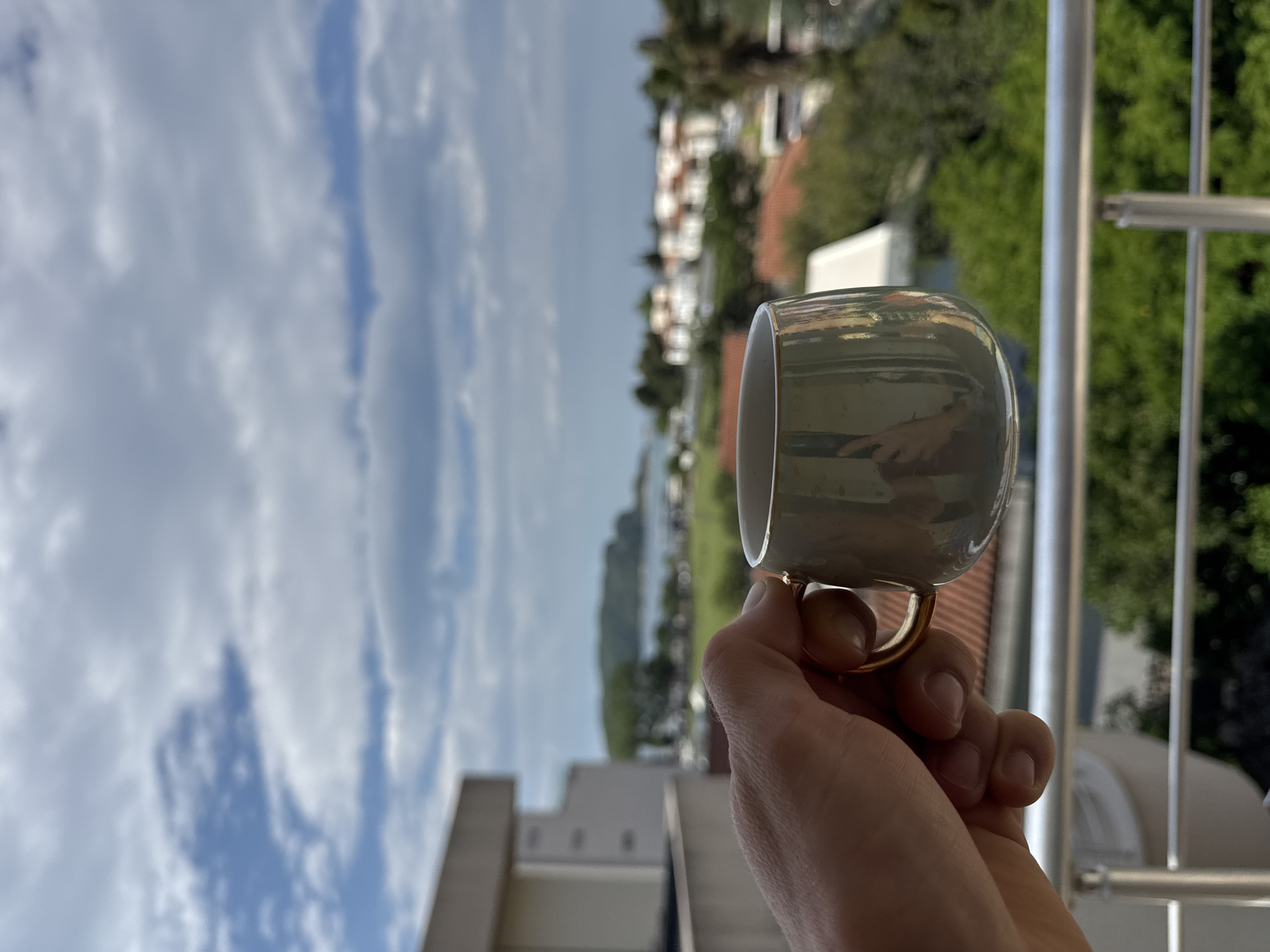 A person holds a light gray mug with a gold handle in front of an urban outdoor scene. The mug is the foreground object, held in the lower right quadrant of the frame. The rest of the frame is filled with an out-of-focus view of a city scene, including buildings, trees, and water in the distance. A clear blue sky with fluffy white clouds is visible above the city. A metal railing is partially visible on the right side of the frame, indicating the scene is likely viewed from a balcony.
The location is likely Budva, Montenegro based on the architecture and landscape.