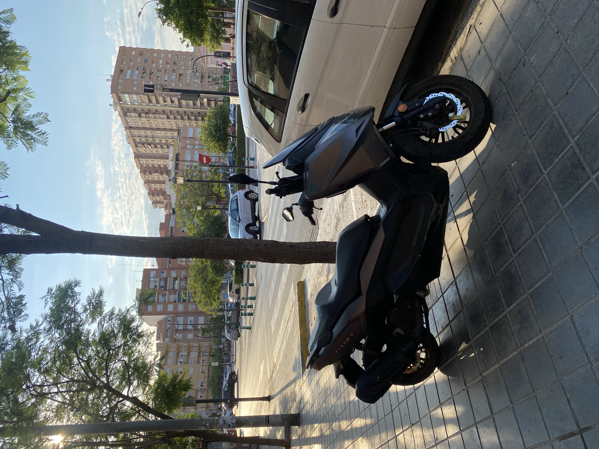 The image captures a street scene in Valencia, Spain, bathed in the warm glow of late afternoon sunlight. In the foreground, a dark gray scooter, identified by the "STORM T" text on its side, is parked on a tiled sidewalk. The scooter has a large windscreen, a black exhaust, and a disc brake visible on the front wheel. To the right of the scooter, a portion of a white vehicle is visible, parked alongside the curb.
The background reveals a city intersection with a pedestrian crossing marked by white stripes. Several cars are present on the road, and green bollards delineate a parking area. In the distance, multi-story apartment buildings with numerous windows line the street, and trees with green foliage provide shade. The sky is a pale blue with scattered white clouds. The overall impression is a typical urban environment in Valencia during a pleasant time of day.
