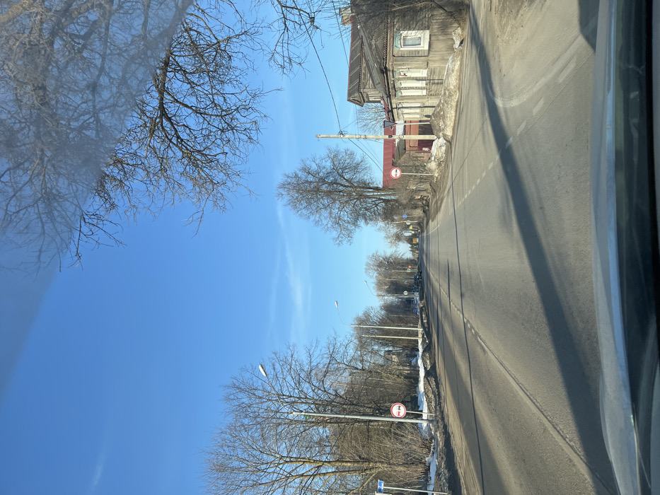 The image depicts a straight, asphalt road extending into the distance under a clear, bright blue sky in Maksimkovo, Russia. The perspective is from inside a vehicle, with the top of the dashboard visible in the bottom foreground. The two-lane road features a broken white centerline and is flanked by shoulders with visible remnants of dirty snow and bare, deciduous trees, suggesting a late winter or early spring setting. Long shadows are cast across the road surface from trees and structures, indicating direct sunlight.
On the left side of the road, multiple bare trees are visible, interspersed with streetlights. A blue street sign is partially visible near the foreground. A circular traffic sign with a red border, depicting two vehicles (one red, one black) side-by-side with a diagonal red line, indicating "No Overtaking," is positioned closer to the road. Further down the road, several parked vehicles are visible on the left shoulder.
On the right side of the road, similar bare trees line the shoulder, along with utility poles and overhead wires. Another "No Overtaking" traffic sign, identical in appearance to the one on the left, is visible. Adjacent to this sign stands a weathered two-story wooden house with multiple windows, some featuring decorative trim, presenting a traditional architectural style. Behind this house, a smaller, red-colored building is partially visible. No individuals or active interactions are visible within the scene.