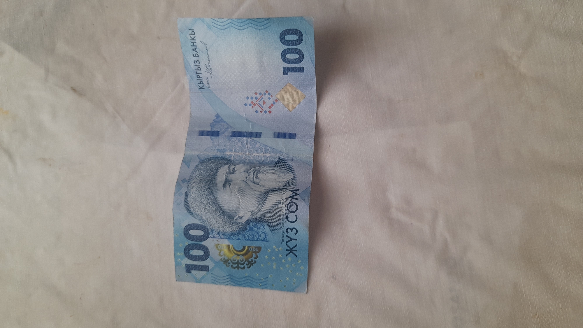 This is a close-up, eye-level, overhead shot of a crumpled, light blue Kyrgyzstani 100 som banknote, placed on a textured, off-white fabric surface. The banknote is oriented horizontally across the frame, slightly off-center to the right. The fabric appears to be a tablecloth or similar material, with visible creases and subtle discoloration in places. The banknote itself features a portrait of Toktogul Satylganov, a Kyrgyz poet and composer, on the left side. The number "100" is prominently displayed in the upper left and lower right corners. The text "КЫРГЫЗ БАНКЫ" (Kyrgyz Bank) is visible in the upper right corner. There are no people present in the image, nor any indication of the city of Osh or Kyrgyzstan other than the text on the banknote itself. The lighting is even, with no harsh shadows.