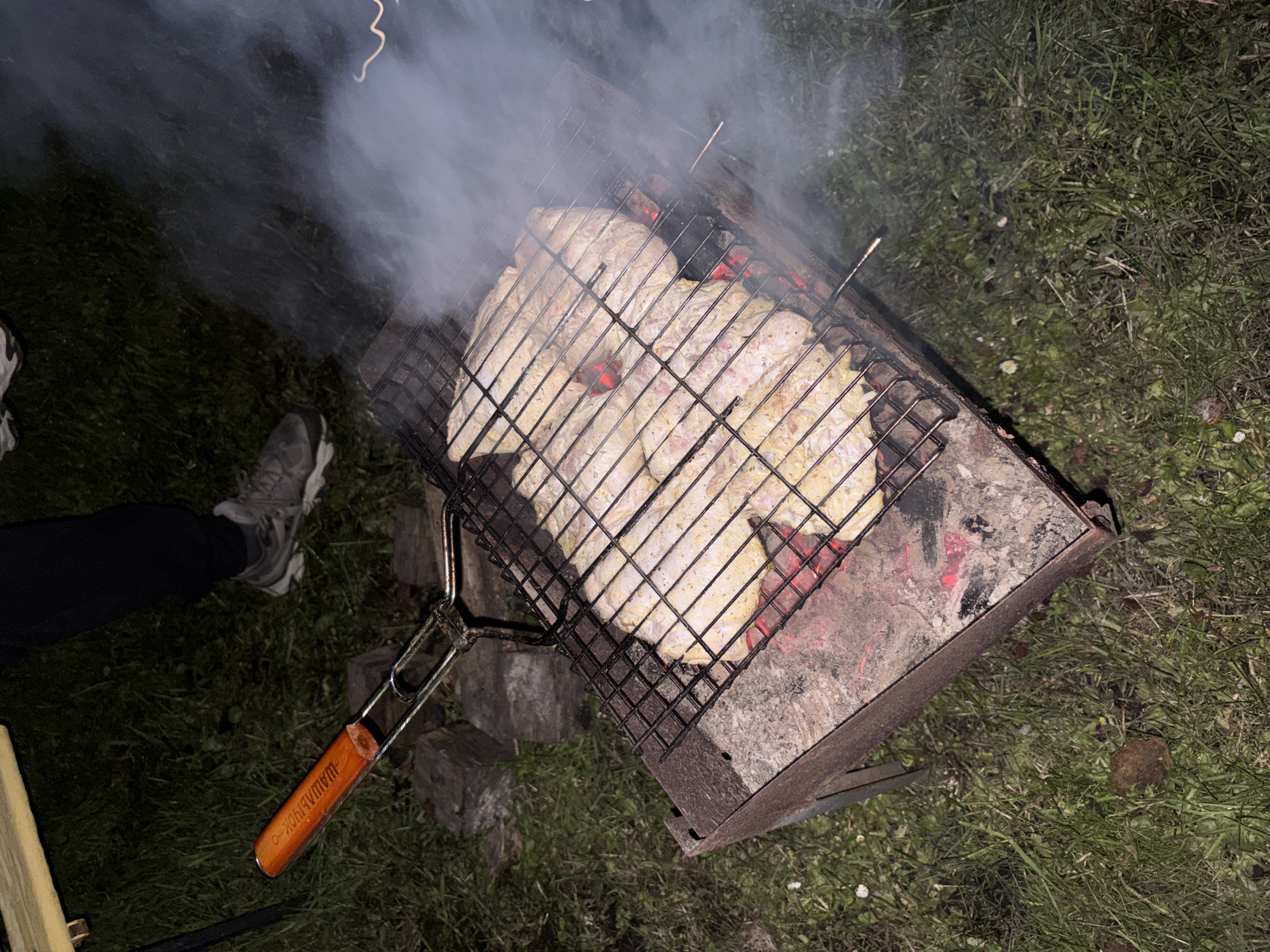 A charcoal grill is set on grass at night, emitting smoke and cooking what appears to be meat. The location is Пярэжырскі сельскі Савет, Belarus. A person wearing sneakers and dark pants stands nearby. The grill is square and open, containing hot coals and a grilling grate with the food. A grilling tool with a wooden handle is visible on the ground near the grill. The grass surrounding the grill is dark.