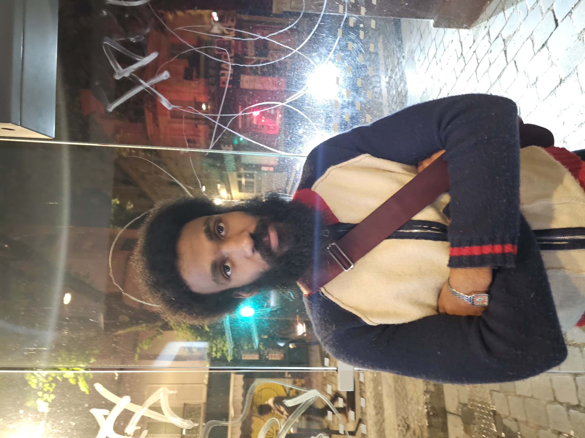 A man with a dark beard and dark hair stands in front of a reflective glass surface, likely a storefront window. He wears a dark blue sweater with red stripes on the cuff and a light-colored vest with a red strap across his chest. A watch is visible on his left wrist. The reflection in the glass shows a blurred urban scene, with lights, buildings, and graffiti. The ground appears to be brick or cobblestone. The environment suggests an urban setting, potentially in Paris, France, given the style of architecture and street design.