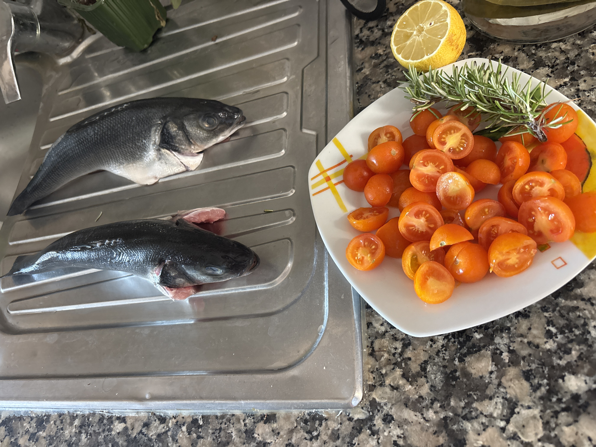 Two whole fish, likely sea bass, are positioned in a stainless steel sink. A bowl of halved cherry tomatoes, a sprig of rosemary, and a lemon wedge are on a speckled granite countertop next to the sink. The scene suggests food preparation in a kitchen. The image does not provide any information about the city of Tavira, Portugal.