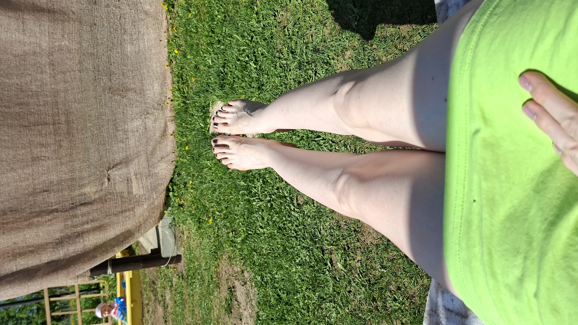 The image presents a view from above, showing a person seated in an outdoor setting. Their legs are stretched out over a patch of green grass, with their feet displaying black nail polish. The person is wearing a bright green garment, and their hand rests on their thigh. A tan-colored canopy or fabric structure is visible to the left. In the background, a partial view of a fence, a yellow structure, and a small child wearing a white hat and blue clothing can be observed. It is daytime, and the scene is illuminated by sunlight. The location is in the city of Большой Букор, Russia.