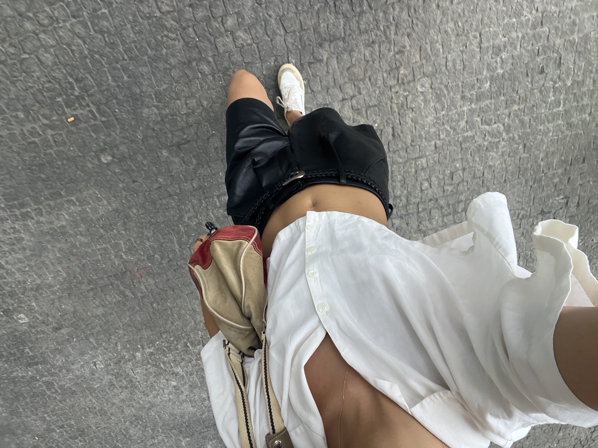 The image shows a person from the chest down, walking on a cobblestone street in Porto, Portugal. The person is wearing a white oversized shirt, black leather shorts, white sneakers, and carrying a tan and red shoulder bag. The cobblestone street is visible in the background, and a cigarette butt is lying on the ground. The lighting suggests it is daytime, possibly late afternoon due to the warm tones. The perspective is from above, looking down at the person.
