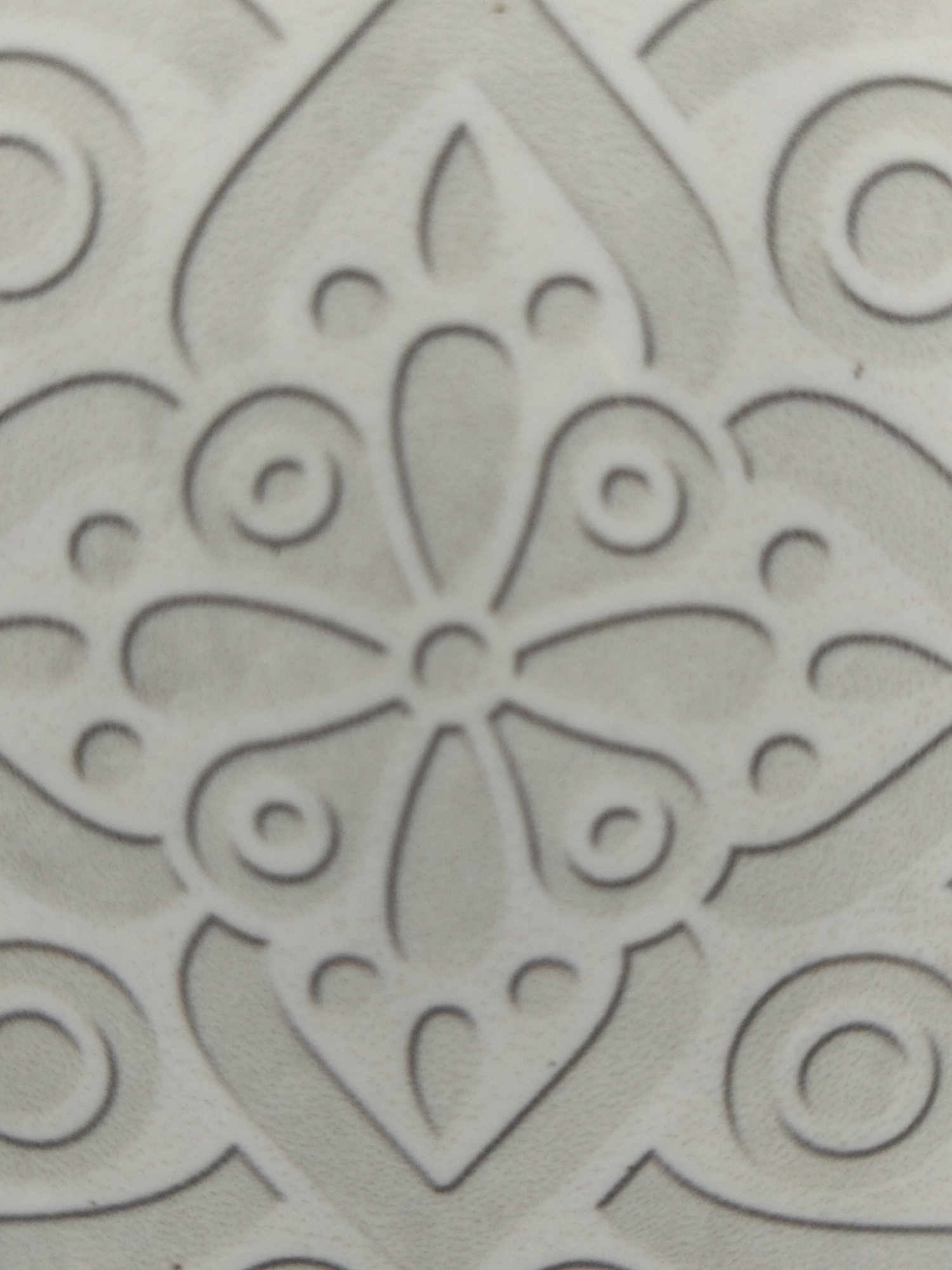 The image is a close-up, high-angle shot of a textured, light-colored surface with an embossed decorative pattern. The pattern features a central floral motif with swirling, paisley-like elements and circular accents. The embossed lines are a darker shade than the background, creating a subtle relief effect. The texture of the surface appears slightly granular. The scene is not identifiable as being located in Kogon City, Uzbekistan, or any other specific urban location. No people, actions, or interactions are visible.