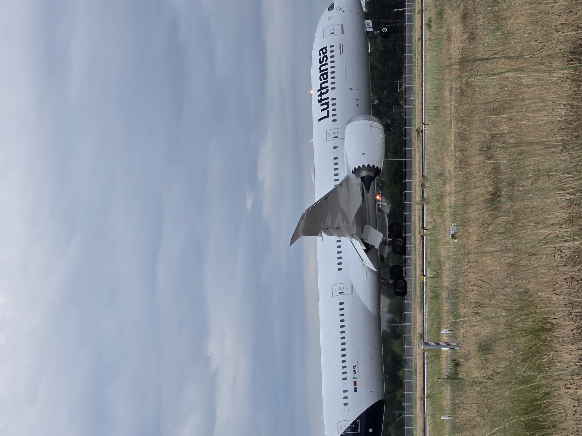 This is a photograph of a Lufthansa Boeing 787-9 aircraft, registered as D-ABPO, in flight near the city of Kelsterbach, Germany. The aircraft is captured in profile, with its landing gear extended and seemingly moments from touching down or having just taken off. The plane's distinctive white fuselage is emblazoned with the "Lufthansa" logo in bold black lettering. The registration number "D-ABPO" is visible on the lower fuselage, along with a German flag decal. The aircraft's wings are swept back, and the engines are clearly visible.
The foreground of the image is dominated by dry, tall grass, suggesting an airport environment or surrounding fields. The background features a line of trees and a chain-link fence, typical of airport perimeters. In the sky, there are dense, overcast clouds, indicating a potentially gloomy or pre-storm weather day. The overall impression is of a large commercial aircraft in the process of landing or taking off at an airport adjacent to a natural landscape.