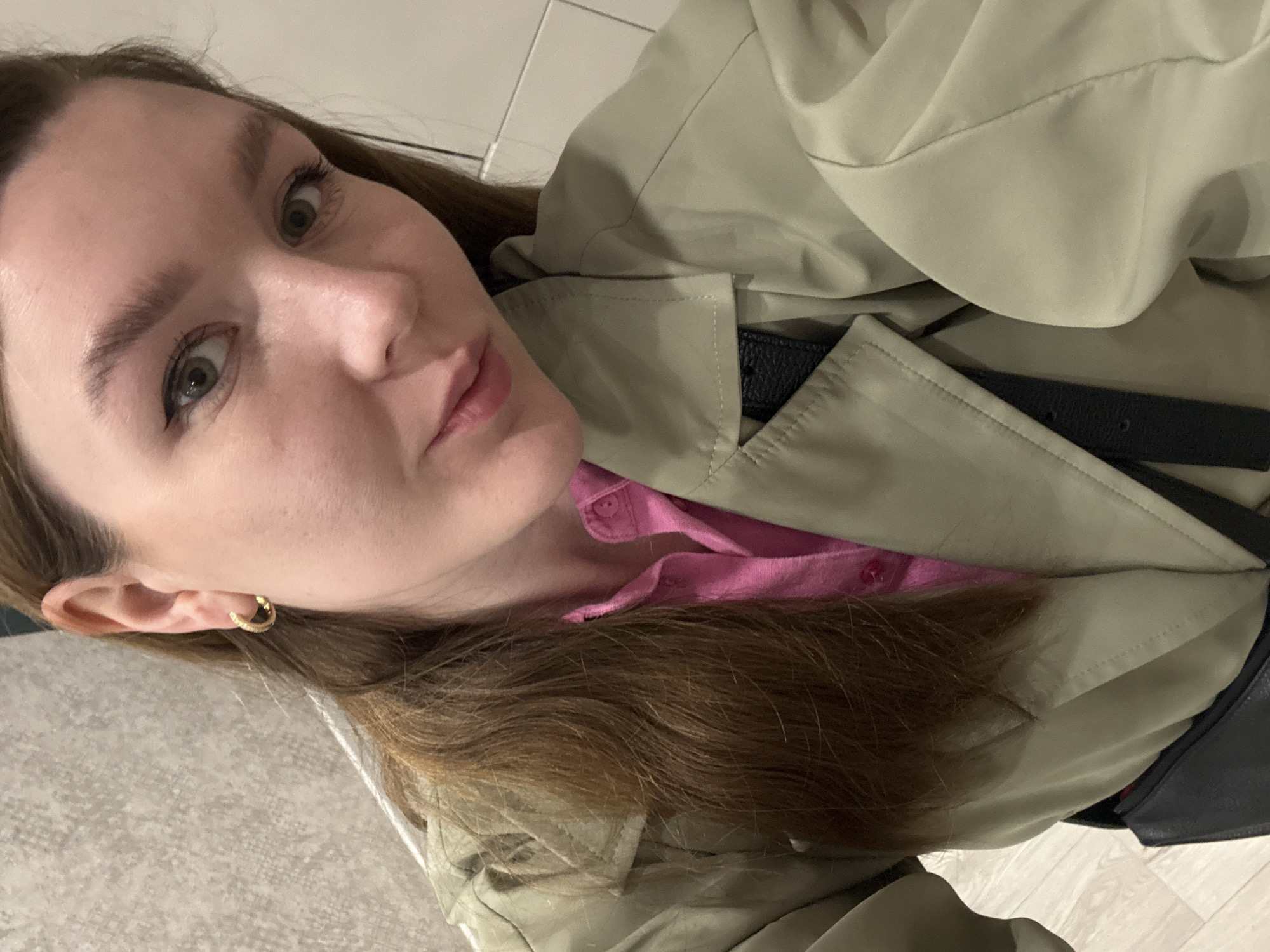 A woman with light skin, green eyes, and brown hair is taking a selfie. She is wearing a pink shirt and a khaki trench coat with a black belt. A gold hoop earring is visible in her left ear. The background appears to be a patterned floor or wall, possibly within an interior space. The location is not identifiable as Izhevsk, Russia based solely on the visual elements present in the image.