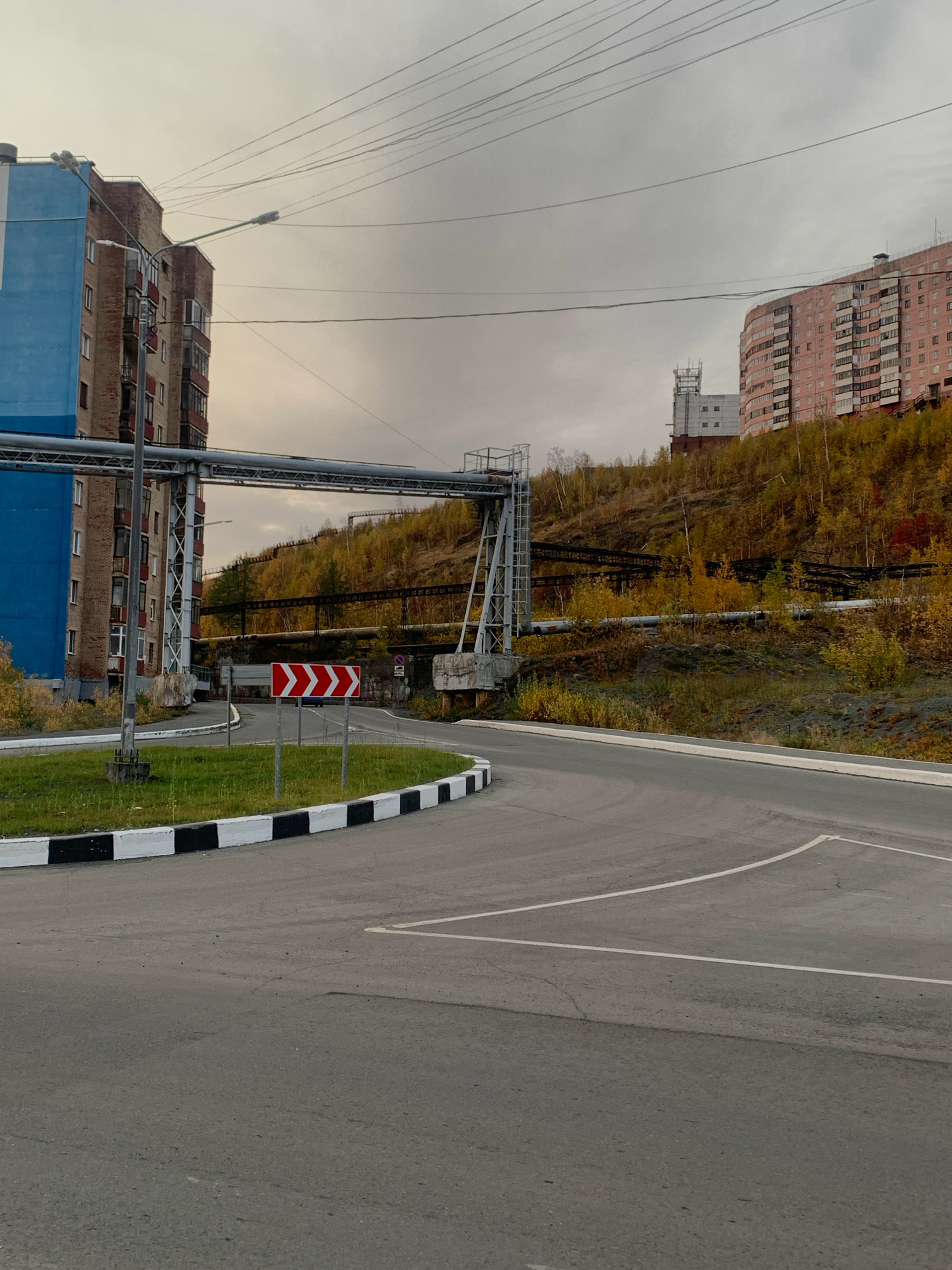 The image depicts an outdoor scene in Norilsk, Russia, likely during autumn. In the foreground, a paved road curves to the right, with white and black painted curbs bordering a grassy island. A directional sign with red chevrons is visible on the island, indicating a curve in the road.
In the middle ground, a blue and brick apartment building stands to the left. Above the road, an industrial pipeline structure spans across the frame, supported by metal beams. The pipeline is elevated and runs up a sloped, wooded hill dotted with yellow and red autumn foliage.
Further up the hill, another brick-colored apartment building is visible, and behind it, a tall, white industrial structure with an antenna. The sky is overcast with a grey, hazy appearance. The overall environment suggests a mix of residential, industrial, and natural elements, characteristic of Norilsk's industrial landscape. There are no people or animals visible in the image.