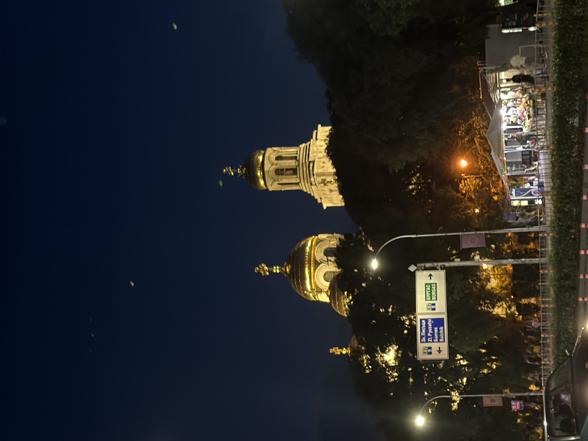 The image captures a nighttime scene in Varna, Bulgaria, dominated by the illuminated domes of the Dormition of the Theotokos Cathedral. The cathedral's golden domes stand out against the dark blue night sky, with bright lights highlighting their architectural details. In the foreground, trees partially obscure the view of the cathedral. To the left, a street sign with Bulgarian text indicates directions to "Zl. Pitsatsi," "Balchik," and "Burgas." Streetlights illuminate the road and a market area with several stalls, one of which displays flowers. Several people are visible near the stalls and walking along the sidewalk. A car is partially visible in the lower left corner of the frame. The overall atmosphere is that of a bustling city at night.