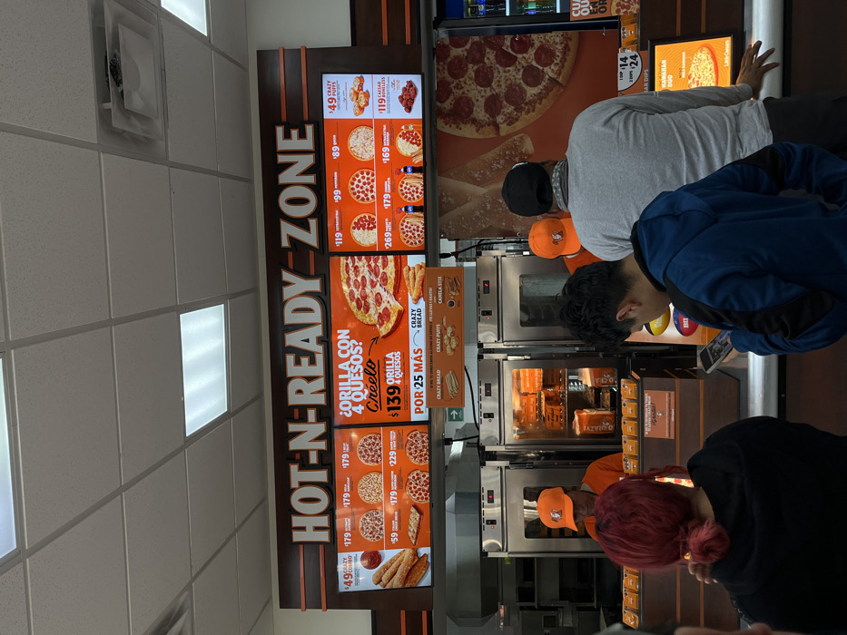 A Little Caesars restaurant displays its menu board labeled "Hot-N-Ready Zone" with prices for various items, including crazy combo, 4 cheese pizza, Caesar wings, and crazy bread. Several people stand in line at the counter, some looking at their phones. Ovens are visible behind the counter. The menu board also shows other options like crazy puffs, canela stix, and dips. The image shows a typical fast food setting at Little Caesars.