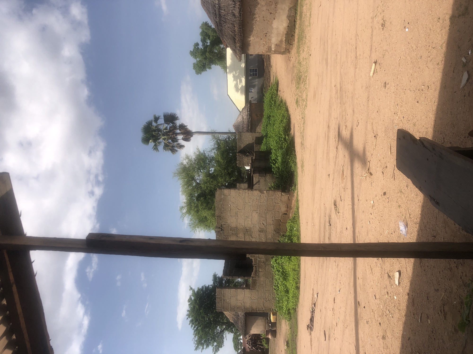 This is an outdoor, eye-level, vertical, full shot taken on a sunny day in Jalingo, Nigeria. The scene depicts a dirt road in front of a collection of buildings. To the left, a partially constructed brick building stands with an open doorway. Adjacent to it is a round structure with a thatched roof, next to which are several earthenware pots and a yellow container. In the center of the image, a tall palm tree reaches towards a blue sky dotted with white clouds. To the right, another round building with a thatched roof is visible, and behind it, a modern building with a light-colored, sloped roof and multiple windows is present. The foreground is dominated by the sandy dirt road, with some scattered debris, including a piece of white plastic and a few sticks. A dark wooden beam, part of an overhead structure, is prominently positioned in the foreground, obscuring a portion of the sky and the landscape.
