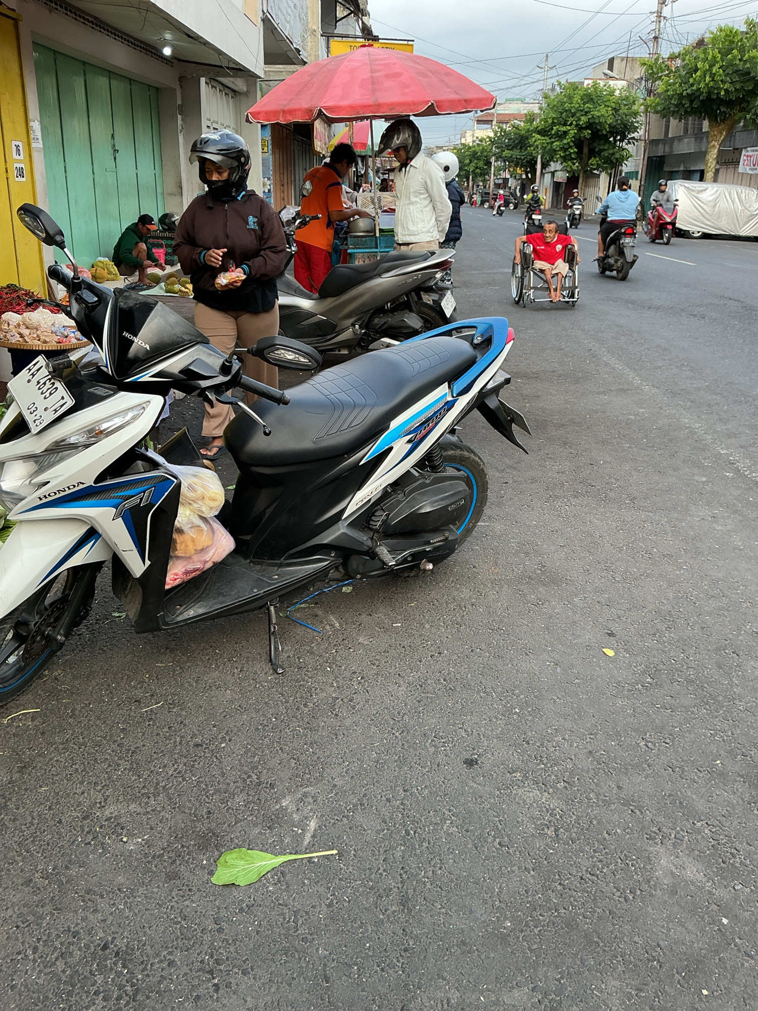 This image depicts a street scene in Magelang, Indonesia, likely in the morning or late afternoon given the lighting. The foreground is dominated by a white and blue Honda motorcycle parked on the asphalt road. Attached to the motorcycle's front is a plastic bag containing what appears to be packaged food items. A person wearing a dark brown jacket and a helmet stands near the motorcycle, holding a small food item.
In the background, several other individuals are present. A person in an orange shirt is seen under a red umbrella, attending to a food stall. Another person in a white jacket is also near the stall. Further down the street, a man in a wheelchair is being pushed by another individual. Several other motorcycles are seen on the road, some being ridden by people. The street is lined with buildings and trees, with power lines visible overhead. A single green leaf lies on the road surface in the lower right portion of the frame. The overall impression is one of a busy, typical street scene in an Indonesian city.