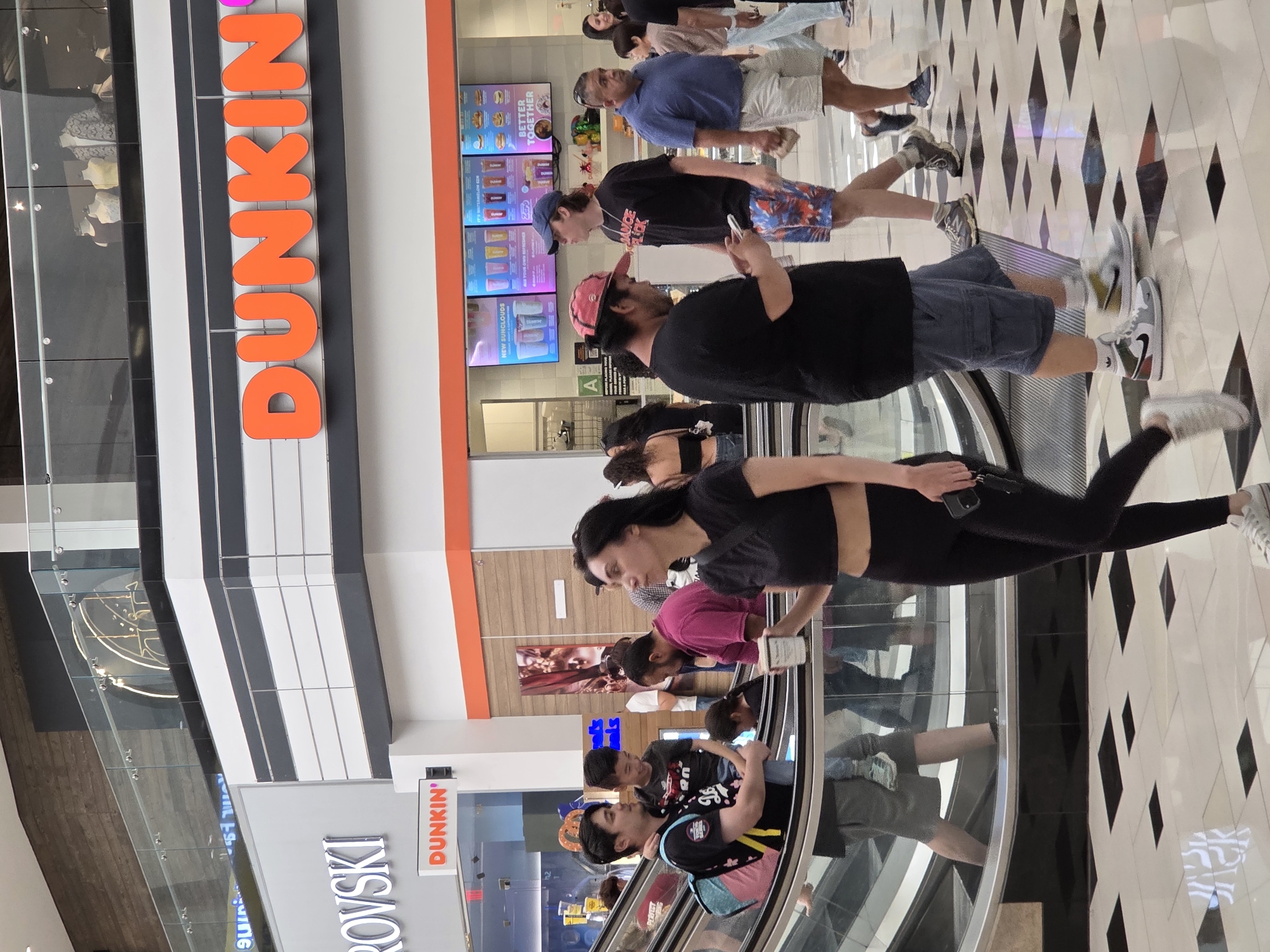 A medium shot captures a bustling scene inside a mall, with an escalator visible on the left. In the foreground, a woman in a black crop top and leggings walks towards the right, holding a coffee cup and her phone. Behind her and slightly to the left, a man in a black t-shirt and shorts is carrying a young boy on his left arm, while another young boy stands next to him. They are positioned on the escalator. Further back, other shoppers are visible, some walking and some on the escalator. A large orange "DUNKIN'" sign is prominently displayed on the upper right side of the frame, indicating the location is a Dunkin' Donuts store within the mall. The mall environment features a modern interior with a tiled floor and large glass railings on the upper levels. The specific location is the Americana at Brand in Glendale, California, United States of America.