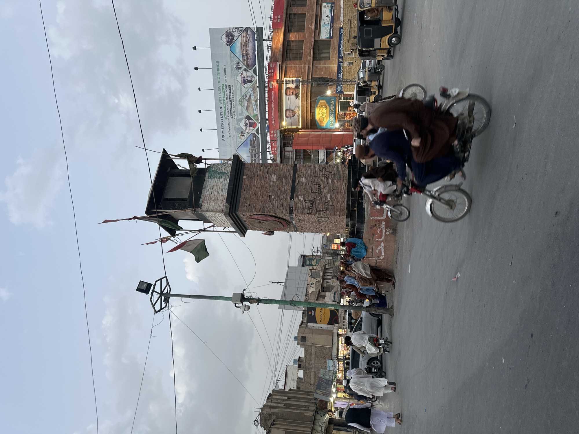 This image shows a street scene in Quetta City Tehsil, Pakistan. The foreground features a brick clock tower decorated with flags. To the right of the tower, people are sitting and walking along the side of the street. In the background, there is a large billboard advertising new commercial plazas and a school, along with a sign for "Quddusi Trading Company." Several auto rickshaws are parked on the street, and motorcycles with multiple riders are in motion. The sky is overcast.