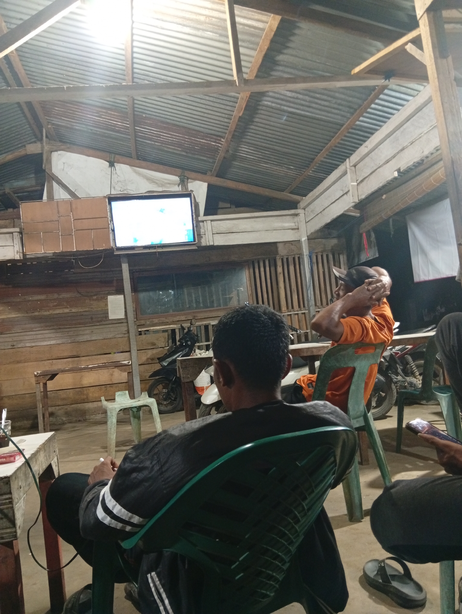 The image depicts an indoor scene, possibly a casual gathering space, with a corrugated metal roof and wooden plank walls. A flat-screen television is mounted on a stand and displays a bright, likely static image. Several individuals are present, seated on plastic chairs. In the foreground, a person wearing a black jacket with white stripes is seated, holding a cigarette. To their right, another person in an orange shirt is visible, wearing a baseball cap and resting their hands on their head. Various objects are scattered around, including a motorcycle parked in the background, tables, and a mobile phone. The overall lighting is dim, with the television screen and ceiling lights providing illumination. The setting appears to be a modest, possibly open-air or semi-enclosed structure, common in informal venues in cities like Lhokseumawe, Indonesia.