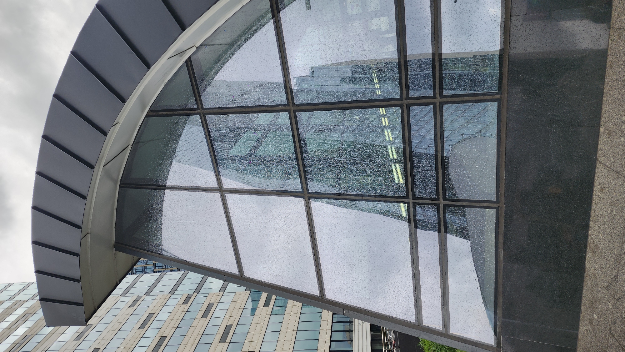 The image depicts a modern architectural detail, likely a covered entrance or passageway, in Moscow, Russia. The structure features a semi-circular roof of dark gray, segmented panels, connected to a frame with large, rectangular glass panes. The glass is covered with raindrops, partially obscuring the view through it. The reflection in the glass shows a modern skyscraper with a grid-like facade of light-colored panels and windows. The right side of the structure is bordered by a dark gray or black granite-like surface. The overall environment is overcast and wet.
