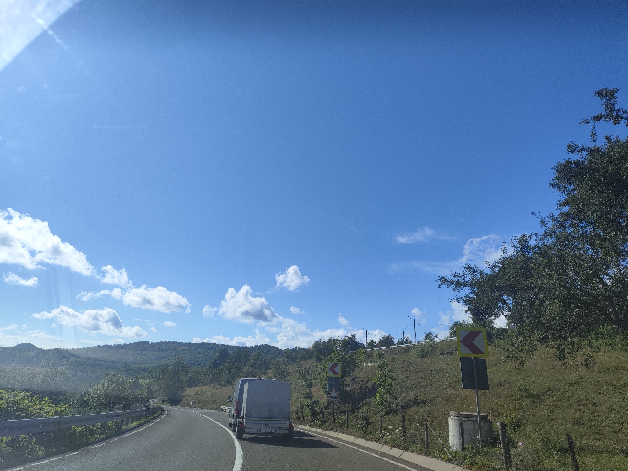 The image captures a view from a vehicle traveling on a paved road in a rural, hilly area. The road curves to the left in the foreground. A light-colored trailer is being towed by a vehicle in the same lane, slightly ahead. To the right of the road, a grassy embankment slopes upwards, dotted with trees and vegetation. Several road signs are visible on the embankment, including a yellow chevron sign indicating a curve to the left and a rectangular sign below it. A concrete structure, possibly a wellhead or culvert, is situated near the base of the embankment. The sky is bright blue with scattered white clouds. The background features rolling hills covered in trees and greenery. There is a guardrail along the left side of the road. The scene suggests a sunny day in a scenic, somewhat remote location, consistent with the landscape that might be found around Petrova, Romania.