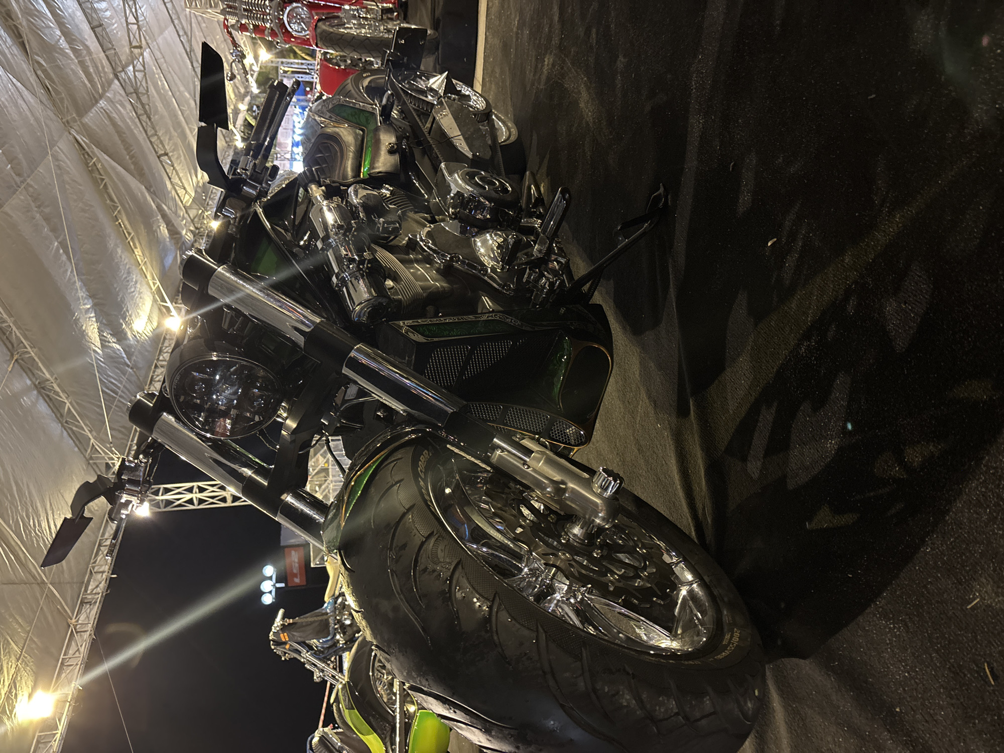 The image features a custom motorcycle prominently displayed, likely at an event or exhibition. The motorcycle is predominantly black with chrome accents and a green highlight on the bodywork. It has a large front tire and a detailed engine with numerous chrome components.
The setting appears to be indoors, underneath a large tent-like structure with a metal frame. The lighting suggests nighttime.
The location is Nha Trang Ward, Vietnam.