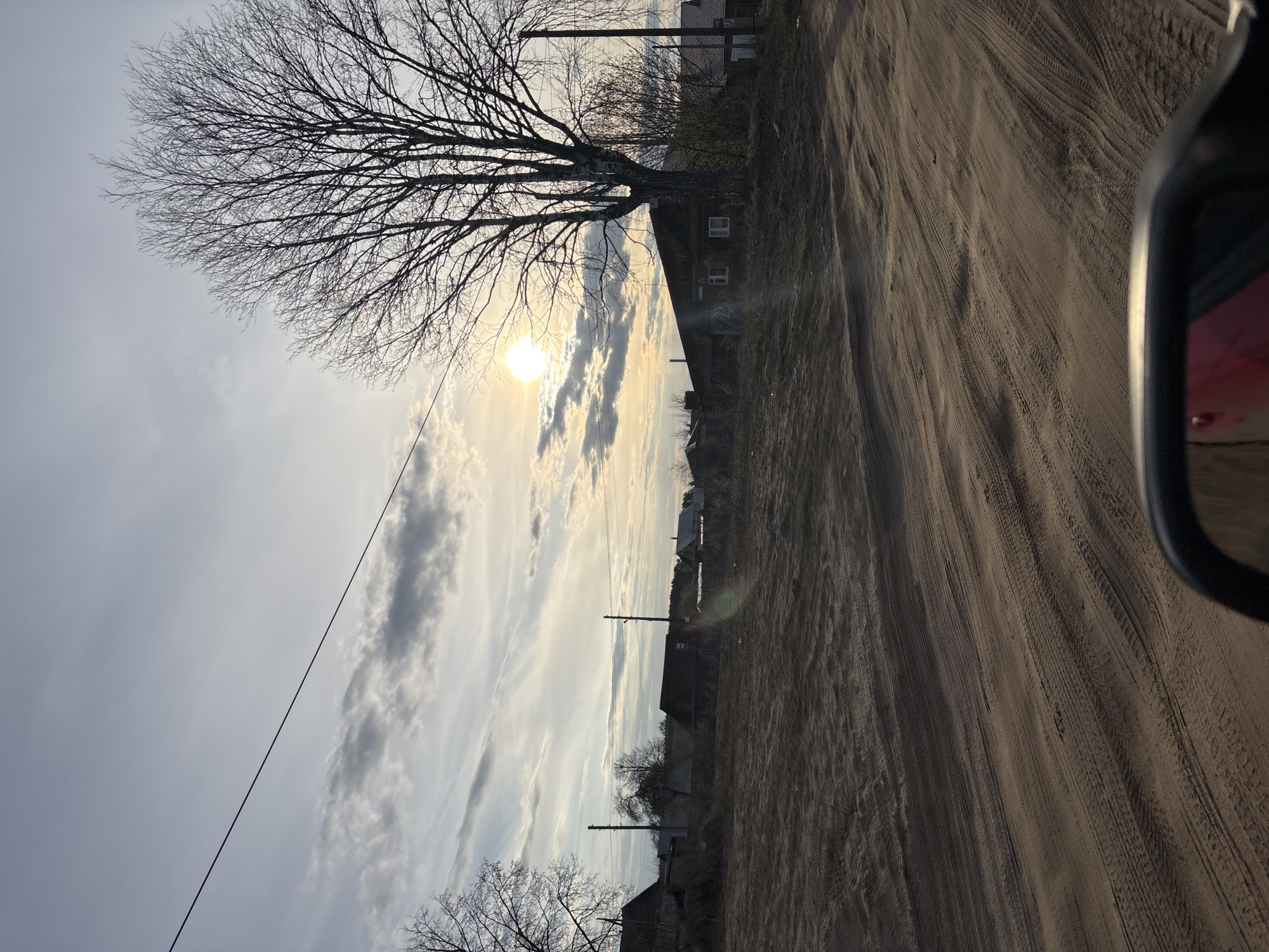 The image depicts a rural dirt road scene, possibly in the vicinity of Chaikovsky, Russia. The road surface is uneven, marked with tire tracks, suggesting vehicular traffic. Adjacent to the road are buildings, possibly houses, with visible windows. A bare tree stands prominently near one of the buildings, indicating it is likely winter or early spring. Overhead, the sky is cloudy with the sun partially obscured, casting a diffused light over the scene. A power line runs across the sky. The image appears to be taken from inside a vehicle, as part of a car's side mirror is visible in the frame.