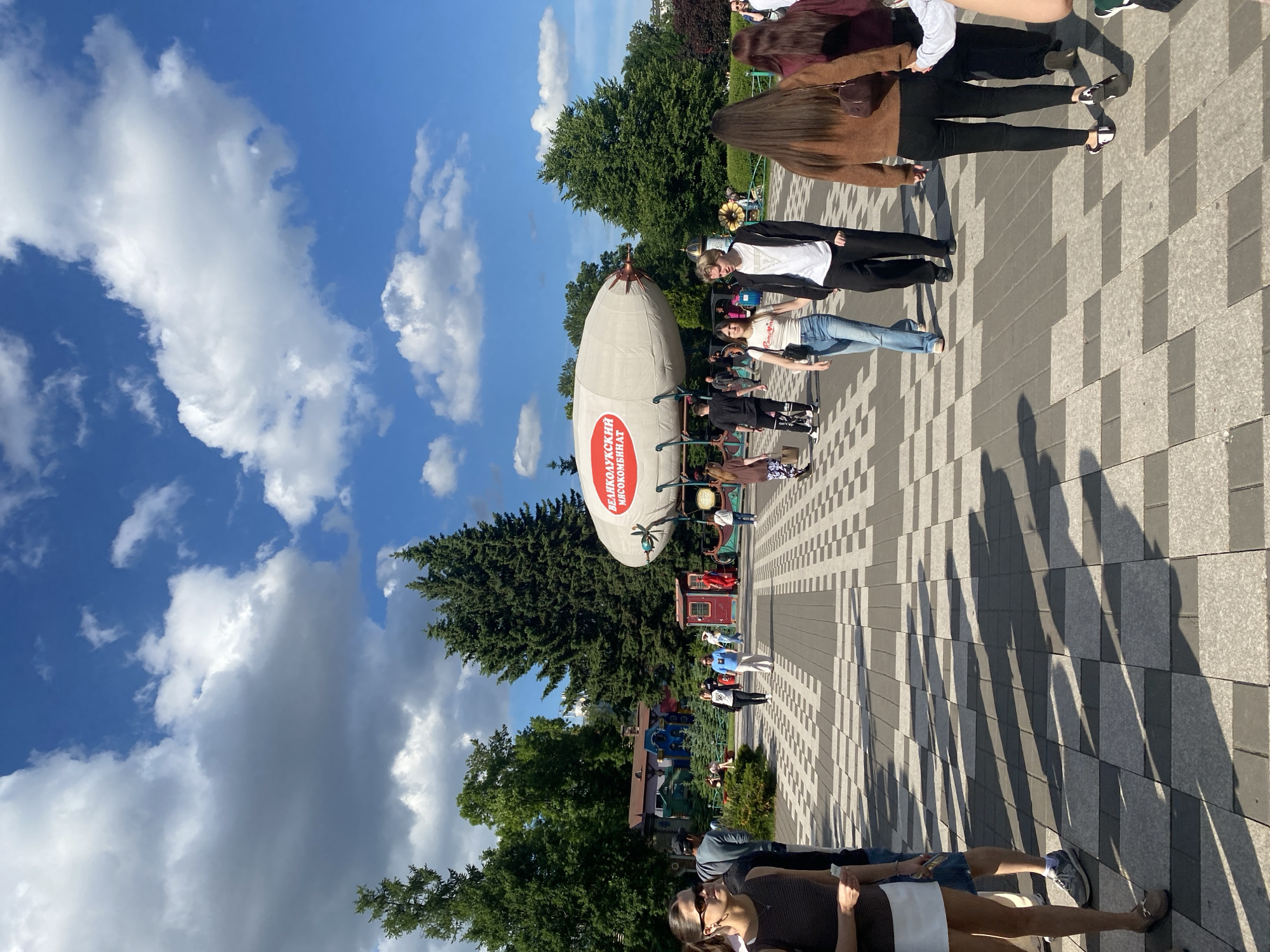 The image depicts a sunny outdoor scene with a checkered paved walkway in a park-like setting. People are walking on the walkway. In the background, a large, light-colored blimp with a red logo is suspended above a structure resembling a train carriage and other colorful buildings. Lush green trees and a bright blue sky with fluffy white clouds complete the environment. The blimp has text in Cyrillic script, which translates to "VEKOLUSSKY MEAT PROCESSING PLANT." The overall atmosphere suggests a public gathering or amusement area.