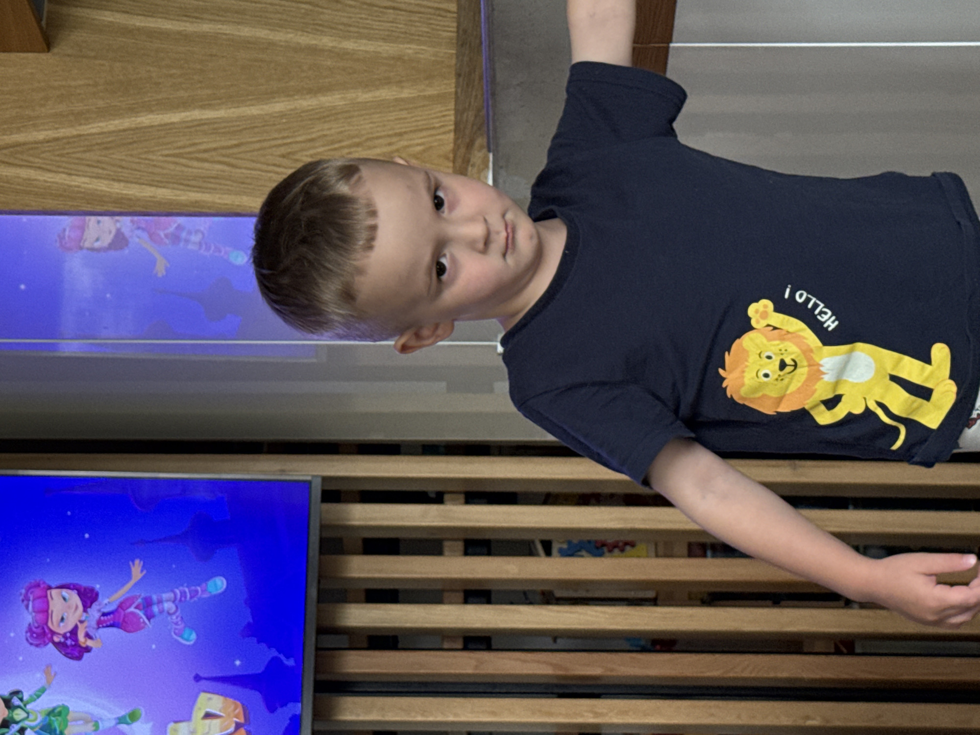 A young boy with short blond hair and a dark blue t-shirt featuring a cartoon lion and the word "HELLO!" is standing in front of a wooden slatted wall. A television screen on the left displays two cartoon characters. The background suggests an indoor domestic setting. The image was taken in Chaikovsky, Russia.