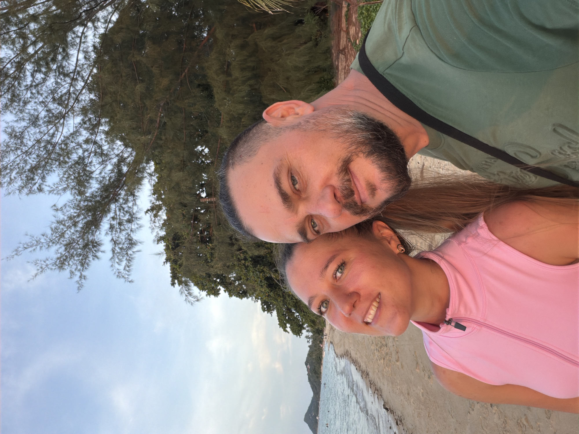 This is a selfie taken on a beach in Baan Thong Po, Thailand. The image features a man and a woman posing for the camera. The woman is in the foreground on the left, smiling. She has blonde hair and is wearing a pink sleeveless top. The man is on the right, closer to the camera, also smiling. He has dark hair, a beard, and is wearing a green t-shirt. Behind them, a sandy beach stretches along the ocean, with gentle waves and a distant shoreline with hills and trees. The sky is a soft blue with some clouds, and lush green trees are visible on the right side of the frame, framing the scene. The overall impression is of a relaxed, scenic beach setting.
