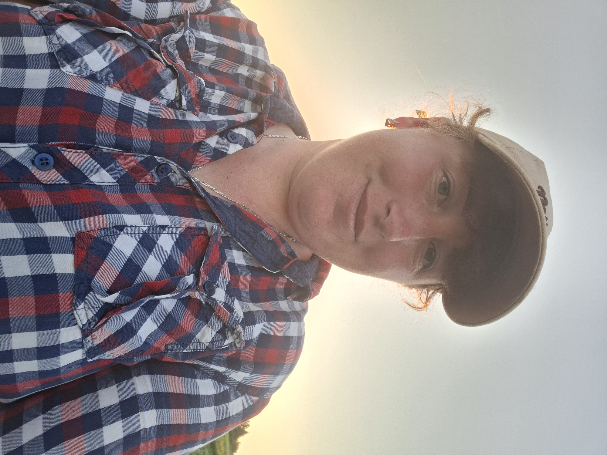 A selfie of a person wearing a beige baseball cap and a red, white, and blue plaid button-down shirt. The person is looking at the camera with a slight smile. The sun is setting in the background, casting a warm glow. The location appears to be outdoors, possibly in a field.