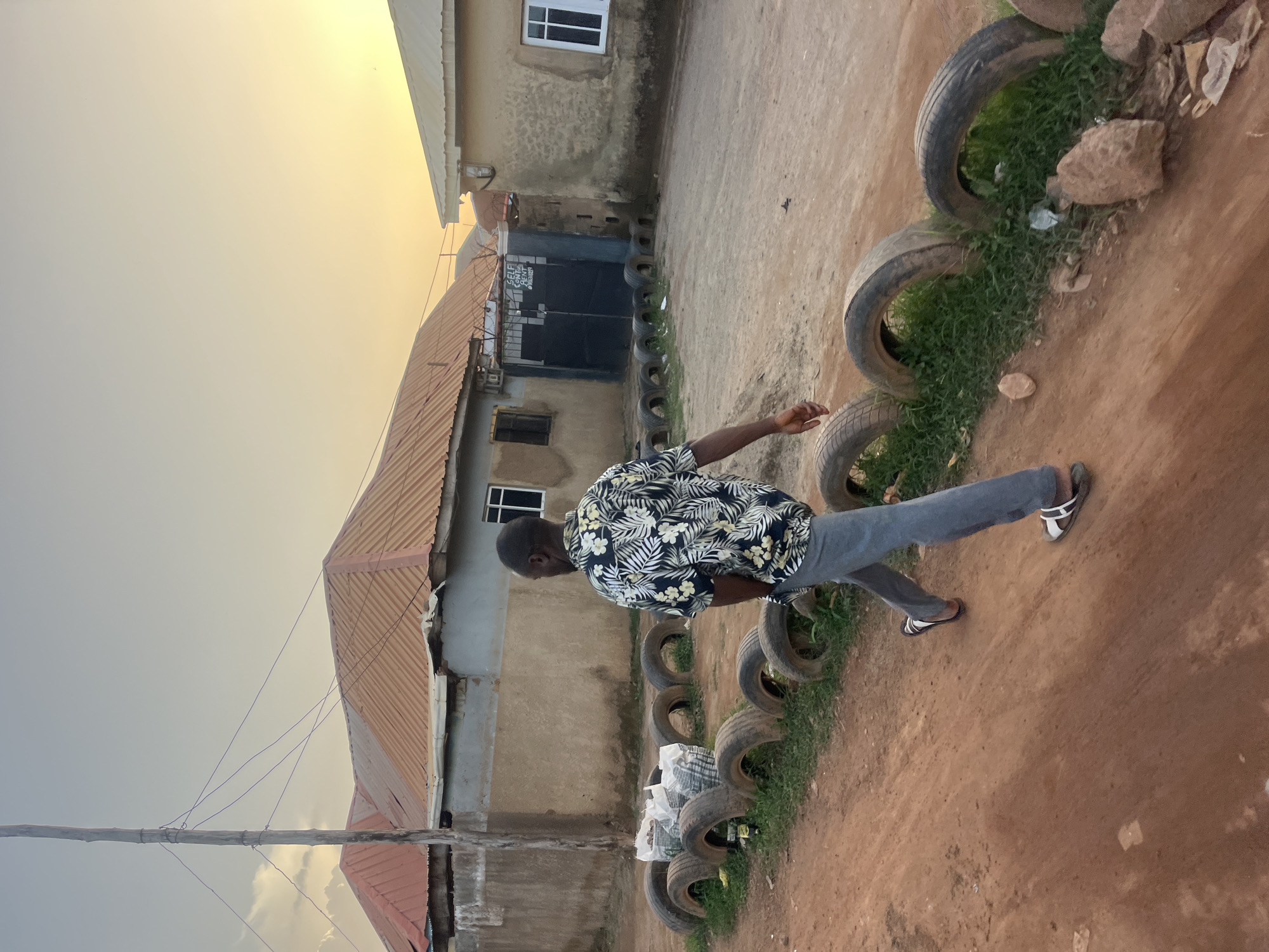A man wearing a floral shirt and jeans is walking on a dirt path alongside a line of old tires filled with grass and some rocks. The path runs alongside a building. In the background, there is a building with a black gate and barred windows. Overhead wires are visible near a pole. The sky is overcast with a yellowish glow. The scene takes place in a residential area. The location is most likely in Abuja, Nigeria due to the style of buildings.