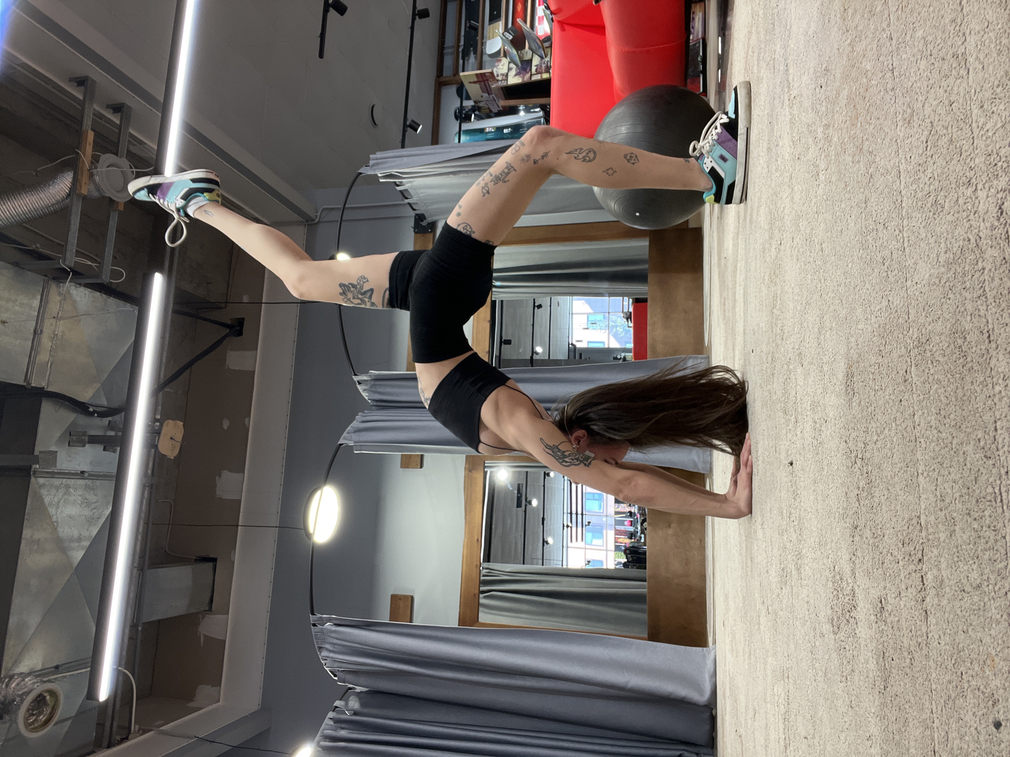 An athletic individual is captured mid-pose, demonstrating impressive strength, balance, and flexibility in what appears to be an indoor gym or studio in Krasnoyarsk, Russia. The person, likely female, is performing an acrobatic handstand or bridge variation. Her hands are firmly planted on the light-colored, textured floor, while her body arches gracefully. One foot rests on a large black exercise ball for support, and the other leg is extended high into the air.
She is dressed in a black sports bra and matching black athletic shorts, complemented by eye-catching multi-colored sneakers that feature shades of turquoise, purple, black, and white. Her long, dark hair cascades downwards towards the floor due to the inverted position. Numerous small, intricate tattoos adorn her legs and arms, depicting various stylized figures, symbols, and possibly flames or clouds. A more elaborate tattoo is visible on her upper right arm.
The setting includes grey pleated curtains, suggesting partitioned areas. The background reveals an industrial aesthetic with exposed ventilation ducts, modern linear fluorescent lighting, and circular light fixtures on the ceiling. Visible in the background are shelves holding various items and a large red object, possibly a piece of gym equipment. A reflection in a mirror or glass partition shows an outdoor scene with buildings, indicating that the activity is taking place during daylight hours. No visible text is present in the image.