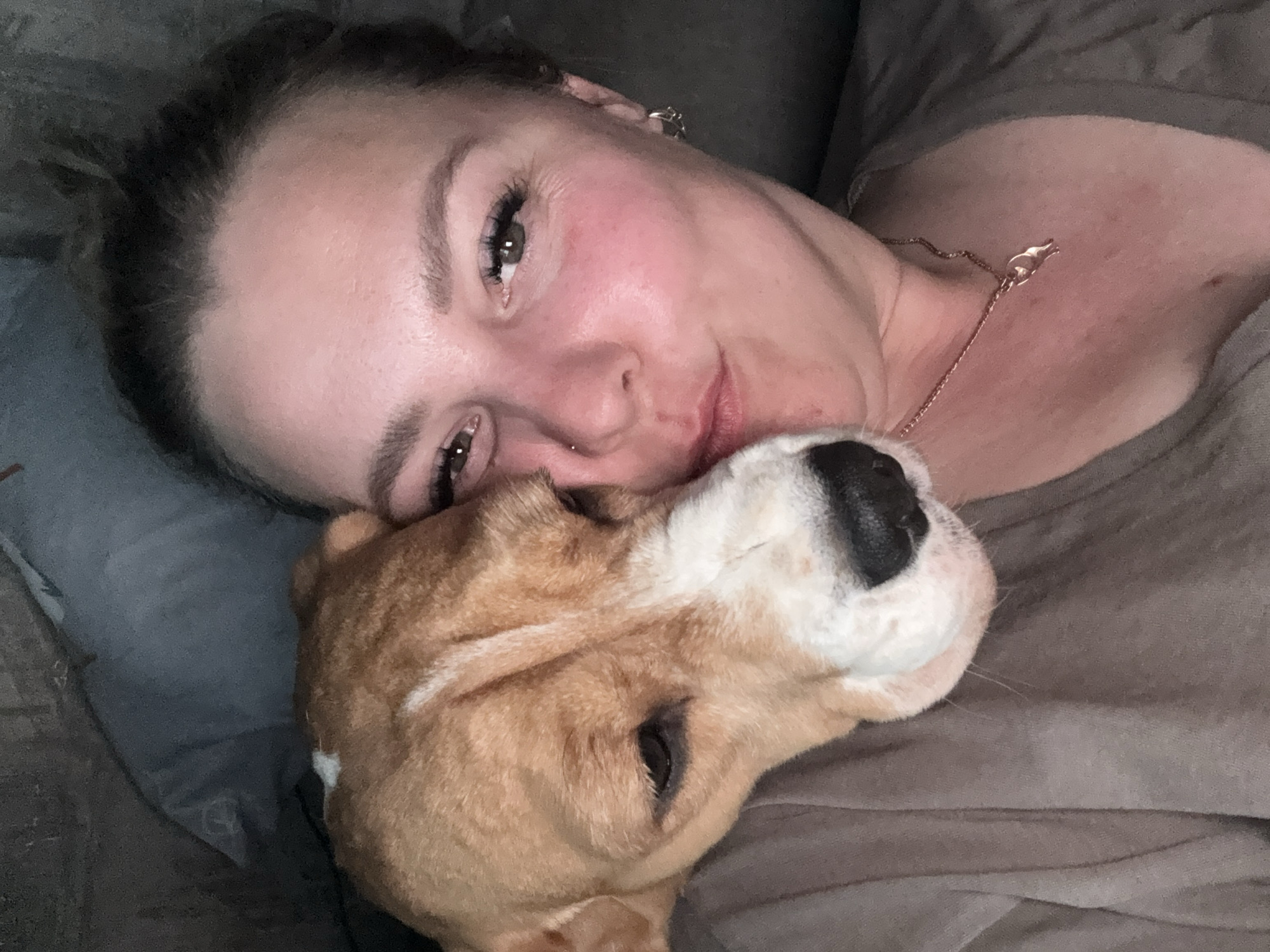 A woman is lying on a gray couch with a tan and white dog cuddled against her face. She has brown hair pulled back, and wears a gold necklace. The location cannot be determined from the image.