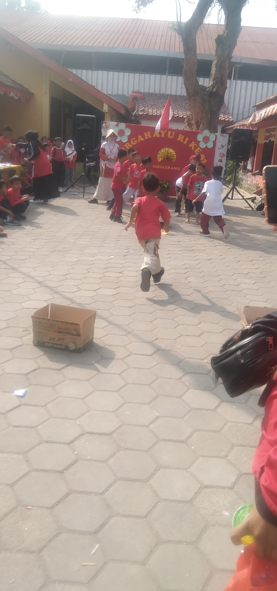 The image shows a group of children in a schoolyard in Rahayu, Indonesia, participating in an event. A banner in the background reads "Hargayu Ri Kesi Cibolerang," suggesting a celebration of Indonesian independence. The children, mostly dressed in red and white, appear to be engaged in an activity or performance. One child in the foreground is running away from the camera, while others are gathered around a woman in a traditional hat, possibly a teacher or organizer. The ground is paved with hexagonal tiles, and a cardboard box sits on the pavement. A tree stands to the right of the banner, and a building with a red roof is visible in the background. The overall scene suggests a festive and communal event for young students.