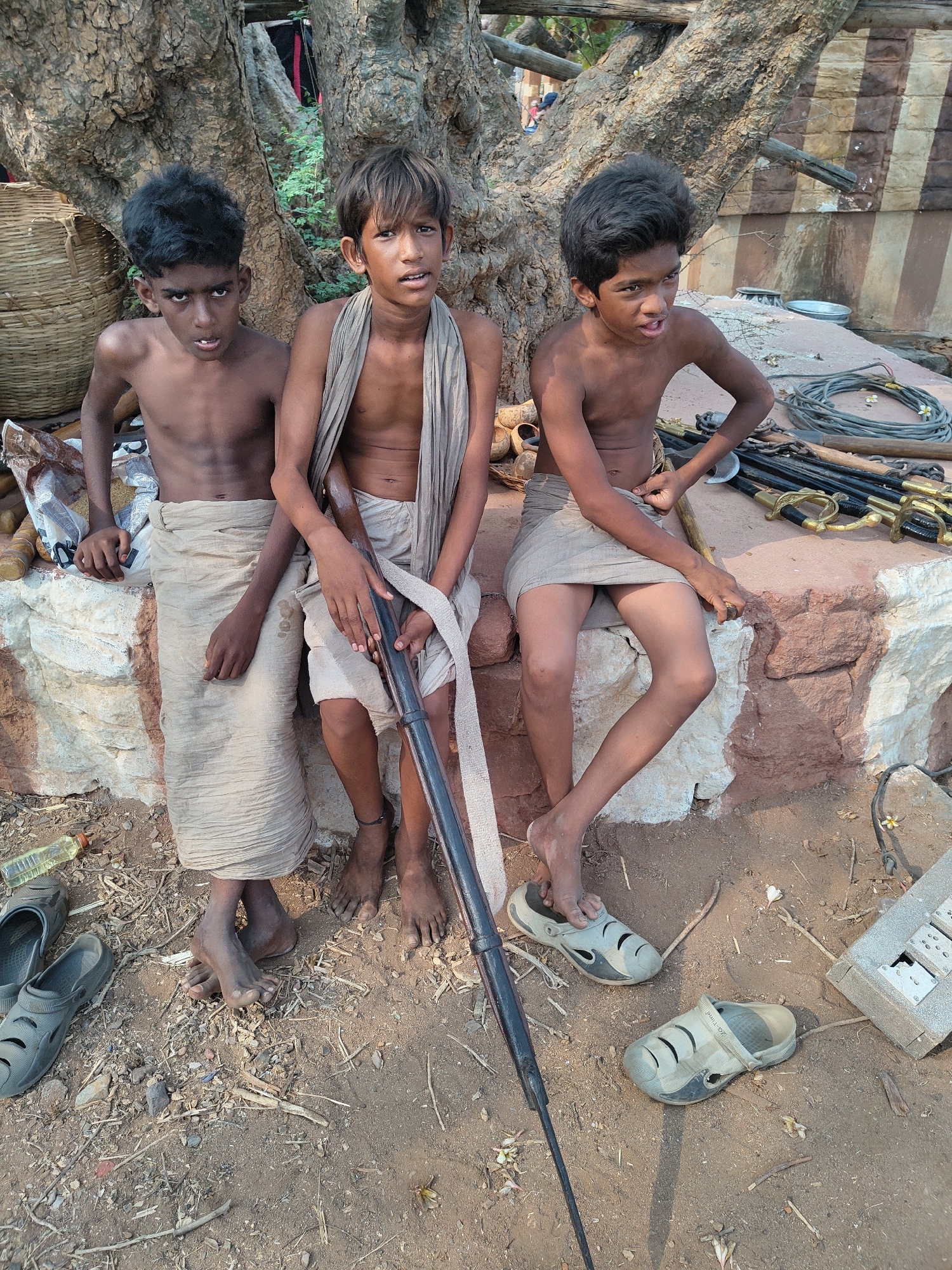 Three young boys are seated on a low stone wall in Dupadu, India. The boy on the left is dark-skinned, bare-chested, and wears a light-colored sarong-like garment. He looks directly at the camera. The middle boy is also bare-chested, wearing a similar garment and a light-colored scarf draped around his neck. He holds a long, dark-colored rod or staff. The boy on the right is bare-chested and wears a light-colored garment. He looks to the right, away from the camera. To the left of the boys is a woven basket and assorted bags or containers. To the right of the boys on the wall are several ceremonial swords with golden handles and coiled wires. On the ground are pairs of sandals, a plastic water bottle, and an electrical box. The backdrop includes the base of a large tree. The ground appears to be dusty.