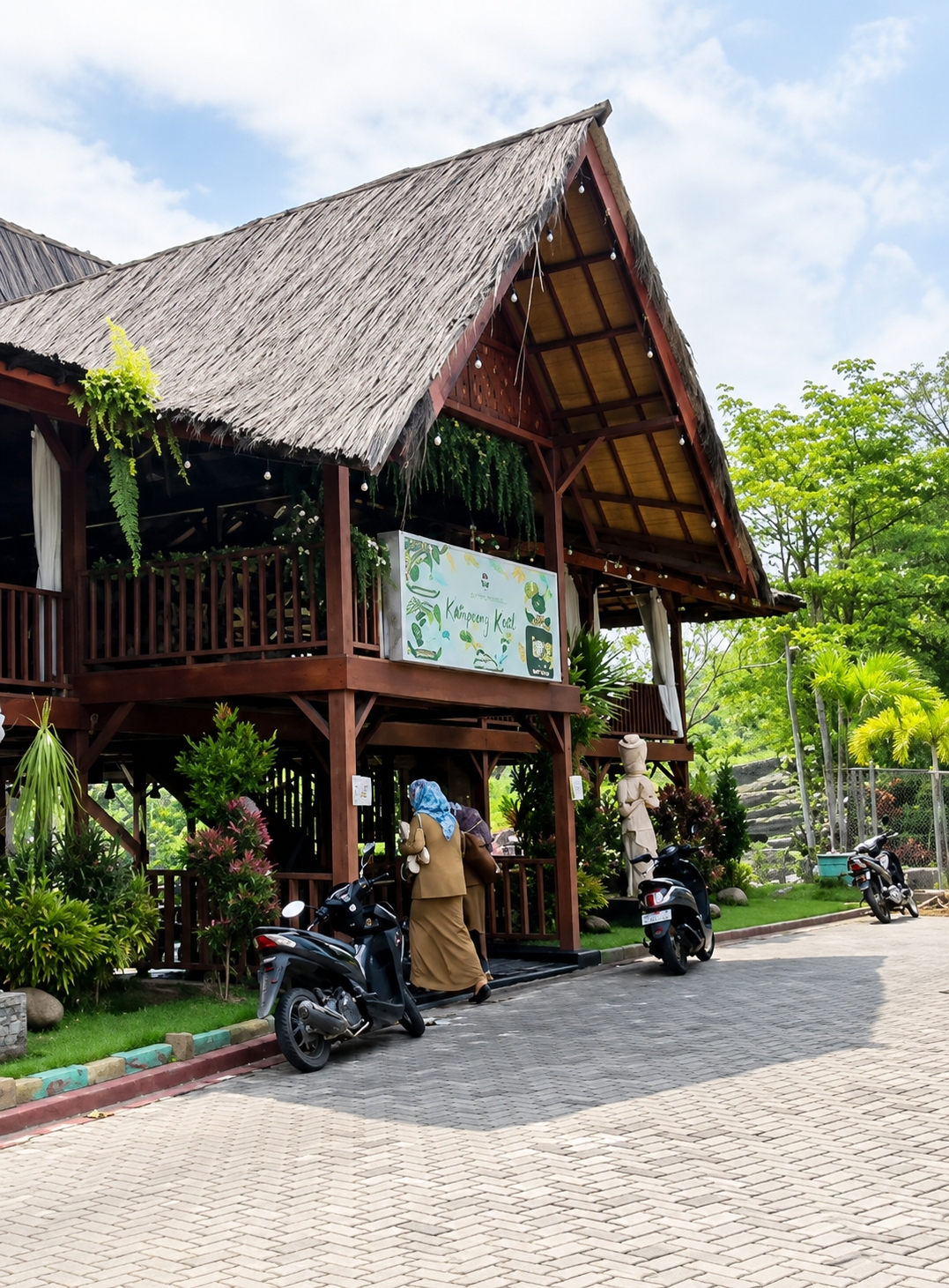 The image depicts a two-story wooden building with a thatched roof, likely a restaurant or cafe, in Eampat Kwalamencirim, Indonesia. The building features open-air levels supported by wooden posts, with balconies and railings. A sign with "Kampong Kecil" is prominently displayed on the upper level. Two individuals, one wearing a tan hijab and abaya and the other in a brown outfit, are seen near the entrance on the lower level. A black motorcycle is parked in front of the building. In the background, lush green trees and vegetation are visible, along with a fence and a stone pathway. The sky is partly cloudy.