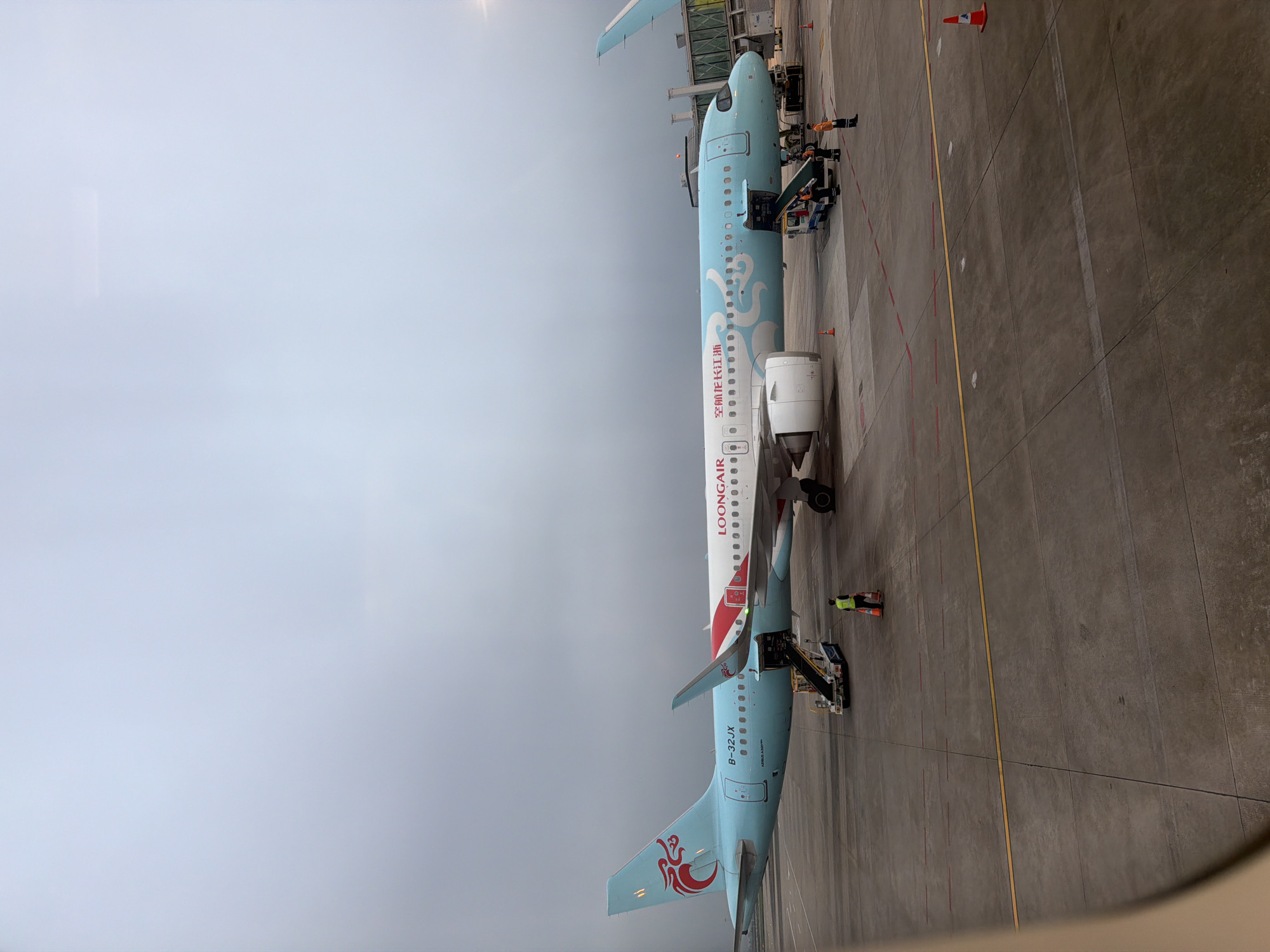 The image shows a Loong Air Airbus A320 aircraft on the tarmac at an airport, likely in Baiyun District, China, based on the airline's operating location. The aircraft has a light blue and white livery with a red and white design near the tail. Ground personnel in high-visibility vests are present near the aircraft, and a boarding stairs are connected to the front door. The ground is marked with yellow and red lines, and orange cones are visible. The sky is overcast.