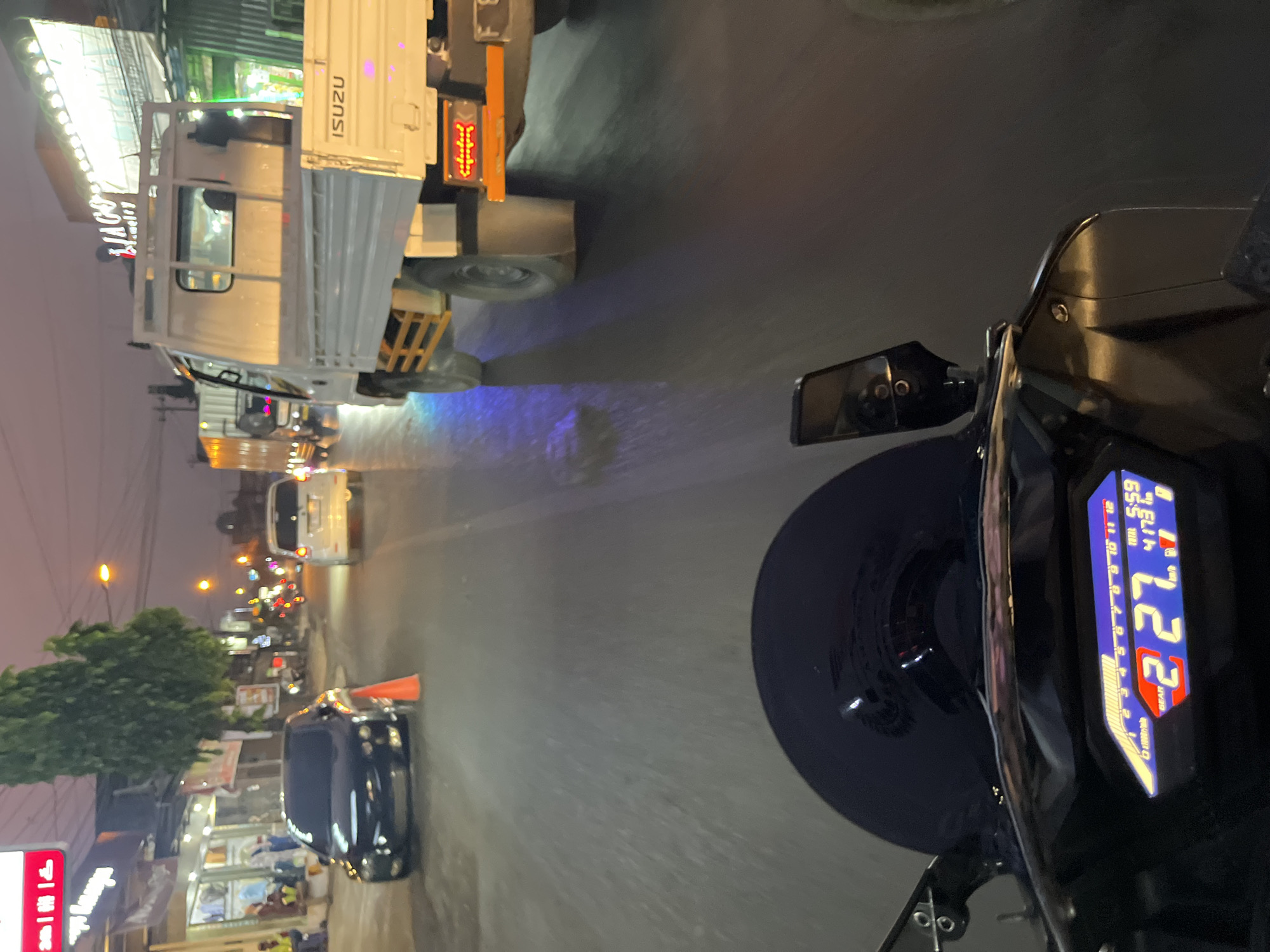This image is a first-person perspective of a motorcycle rider in Kriyan, Indonesia, captured at dusk. The rider's dashboard displays "GEAR 2", a speed of "227", a time of "5:59", and "4173" kilometers. The road is wet, indicating recent rain, and reflects the ambient light from streetlights and vehicle headlights.
In the foreground, the motorcycle's dashboard and windshield are visible. Ahead, several vehicles are present on the road. A black car is parked on the left side of the road, partially obscured by an orange traffic cone. Further ahead, a white van and a white Isuzu truck are traveling in the same direction as the motorcycle. The truck has "ISUZU" branding visible on its side.
On the left side of the road, there are buildings with illuminated signage, including a store with the word "Jewelry" prominently displayed. People are seen near the shops. Streetlights are illuminating the scene, casting a dim glow consistent with twilight. The overall environment suggests a commercial street in a town or city.