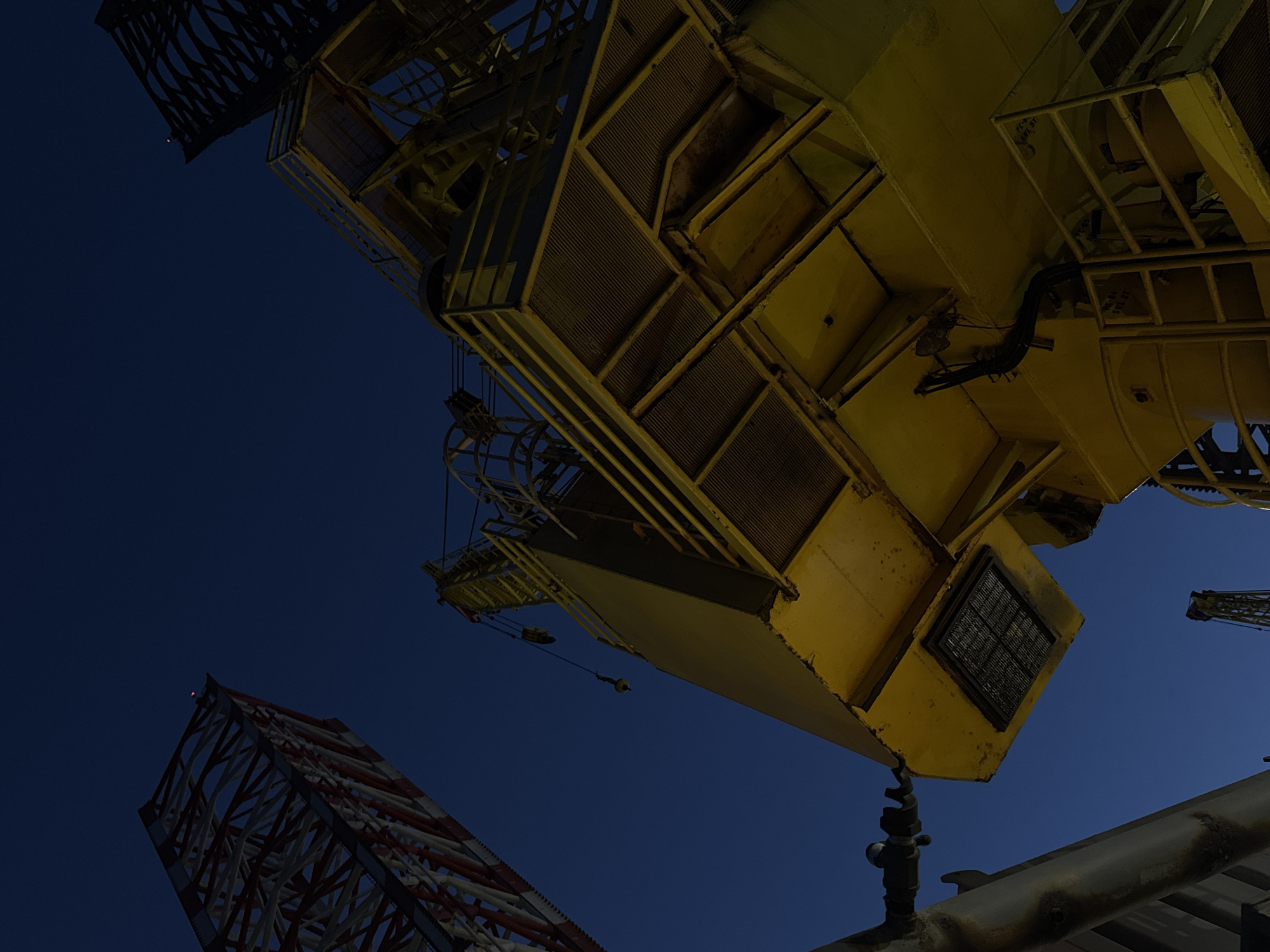 In the foreground, a large yellow industrial structure, likely part of a crane or offshore platform, dominates the frame. Its metallic surfaces are illuminated by a warm light, revealing complex mechanical components, railings, and grates. The structure extends upwards, suggesting significant height. In the background, a tall, lattice-like red and white structure is visible against a deep blue twilight sky. Another similar, darker lattice structure is partially visible to the right. The scene appears to be an industrial or port area, possibly at dusk or night, with minimal visible human activity. The overall atmosphere is industrial and somewhat stark. The location is identified as Unknown, Italy.