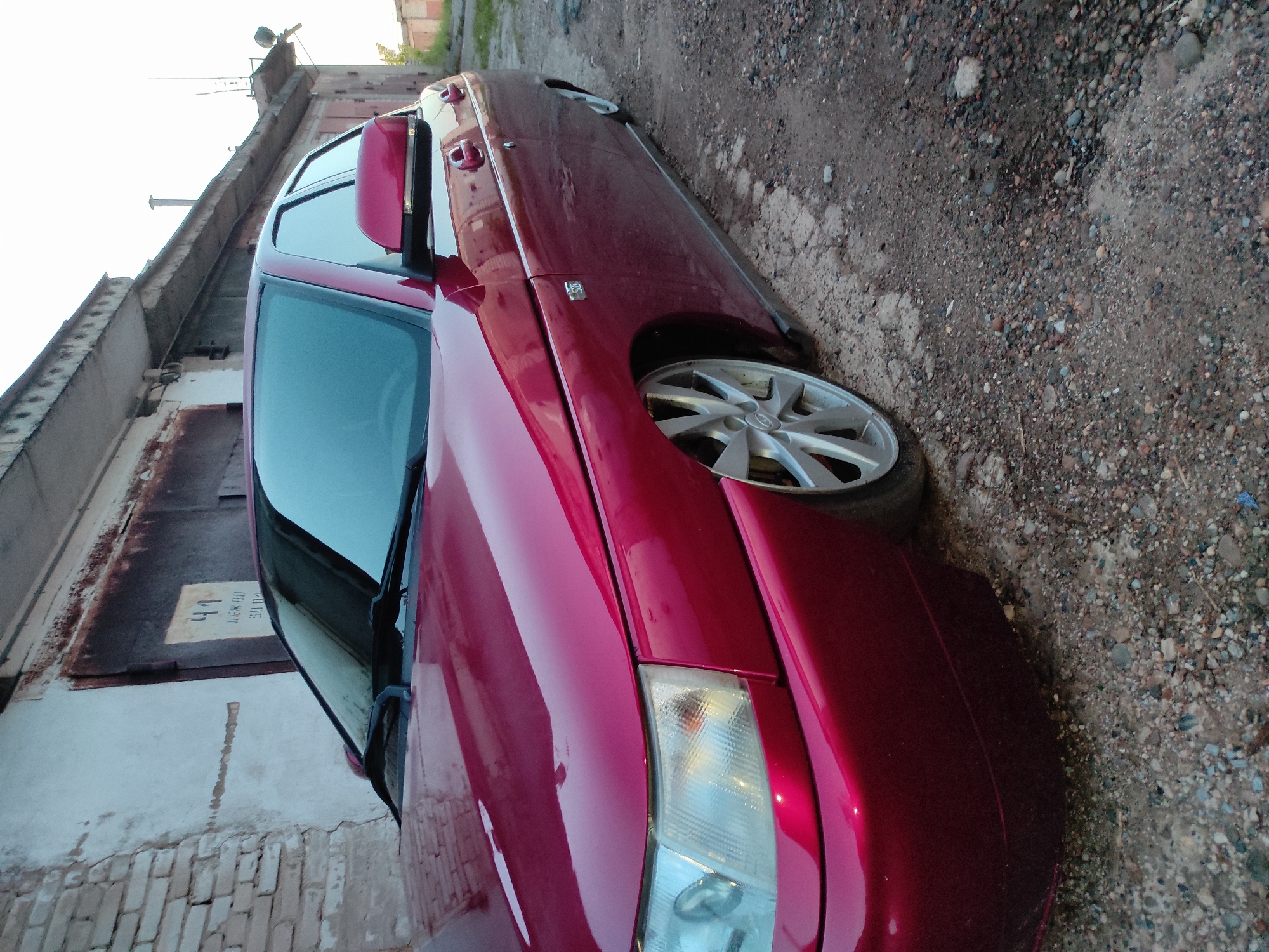 A reddish-maroon four-door sedan is prominently featured, parked on unpaved ground composed of dirt and various sizes of gravel and small stones. The car's metallic paint reflects ambient light. Key visible components of the vehicle include a front headlight, a multi-spoke silver alloy wheel, the front fender, the driver's side door with its handle, and the side mirror. A small, indistinct badge is visible on the front fender above the wheel arch.
The car is situated alongside a weathered building, likely a garage or a series of garages. This structure features a rusty metal garage door. Above and below the door, the wall is made of light-colored plaster or concrete, with visible brickwork at the very base. A concrete or stone ledge runs along the top of the building, where some pipes and what appears to be an antenna are visible against a bright, clear sky. The overall setting suggests an older, possibly industrial or residential area with an unpaved access road or alleyway.
The lighting indicates it is daytime with clear weather. No people are present, and no specific activity is taking place; the car is stationary.
On a white rectangular label affixed to the garage door, oriented sideways, the following Cyrillic text is visible:
- "41"
- "Бл. 04" (likely "Block 04" or similar)
- "30.01" (potentially "January 30th" or an identification code)