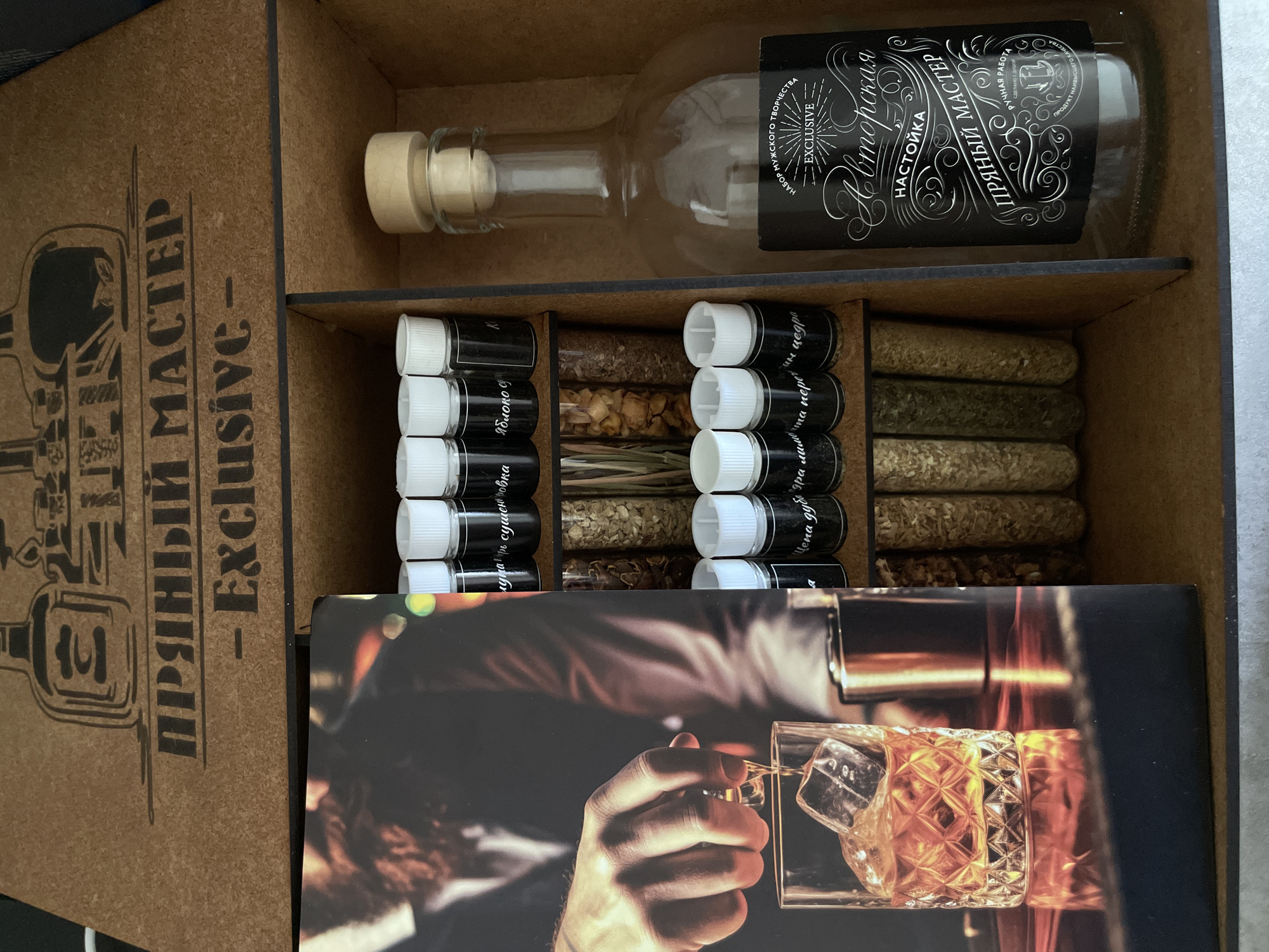 The image depicts a wooden box containing a kit for making infused vodka. The box is divided into compartments, with a bottle lying horizontally in the top section. The bottle has a cork stopper and a black label with white Cyrillic text, including the words "Авторская Настойка" (Author's Infusion) and "Пряный Мастер" (Spiced Master). Below the bottle are compartments containing small vials filled with various herbs and spices, each with a black label and white text. The labels seem to specify the ingredients, such as "Яблоко Суш" (Dried Apple), "Цедра дуба" (Oak Peel), "Рома перца" (Rum Pepper), and "Луна Кир" (Moon Kir). In the lower section of the box, a card displays a photograph of a hand pouring whiskey into a glass with ice cubes. The text on the box side repeats "Пряный Мастер - Exclusive," and the labels on the bottles indicate that the kit is a product of "Ручная работа" (Handmade) and made with "душой" (soul), implying artisanal production. The kit is designed for creating flavored vodka using a variety of natural ingredients.