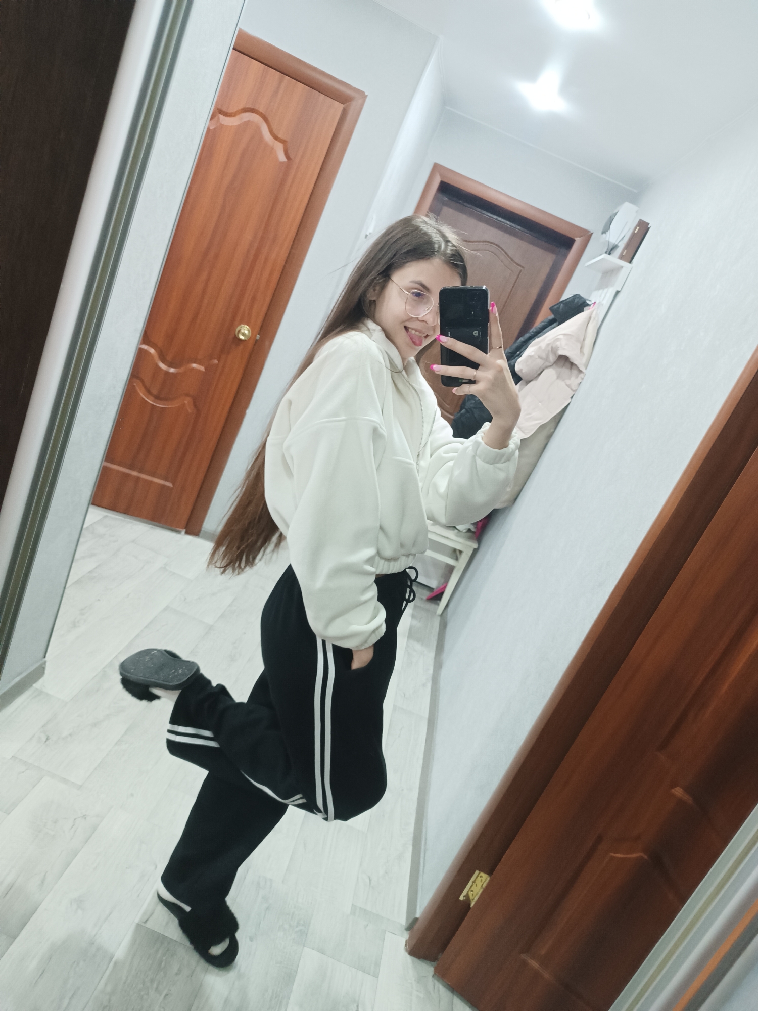 Location: Unknown, Unknown.
Scene: A female individual is depicted indoors, taking a mirror selfie within what appears to be a residential hallway or room. The subject has long, straight dark brown hair and wears round, clear-framed glasses. She is dressed in a white, long-sleeved cropped hoodie, black sweatpants with two vertical white stripes on the outer seams, and black furry slip-on slippers. Her fingernails are painted pink, and she wears rings on multiple fingers. She holds a black smartphone in her right hand, pointed towards the full-length mirror. Her expression is a smile with her tongue slightly extended. Her right leg is lifted and bent backward at the knee, while her left hand is placed in her pocket. The immediate environment consists of light gray walls and light-colored, possibly laminate, flooring. Two wooden doors are visible: one partially obscured dark brown door to the left, and another medium brown door with a gold spherical doorknob and two decorative wave-like carvings. Overhead lighting fixtures are reflected in the upper portion of the mirror. A white wall shelf containing unidentified objects and a light-colored jacket draped over a fixture are discernible in the reflection behind the subject.