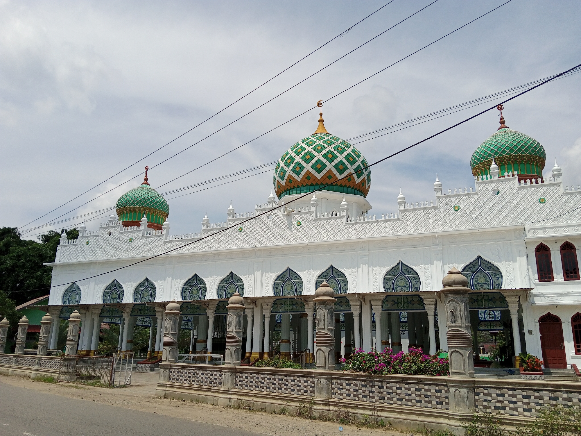 The image shows a white mosque with multiple green and gold domes, located in Pulorayeuk, Indonesia. The mosque has a long, colonnaded porch with decorative blue and white arched windows above. In front of the mosque is a decorative fence and a paved area that appears to be a road or pathway. Several power lines are visible crossing the sky above the mosque. There are no people visible in the image. The sky is overcast.