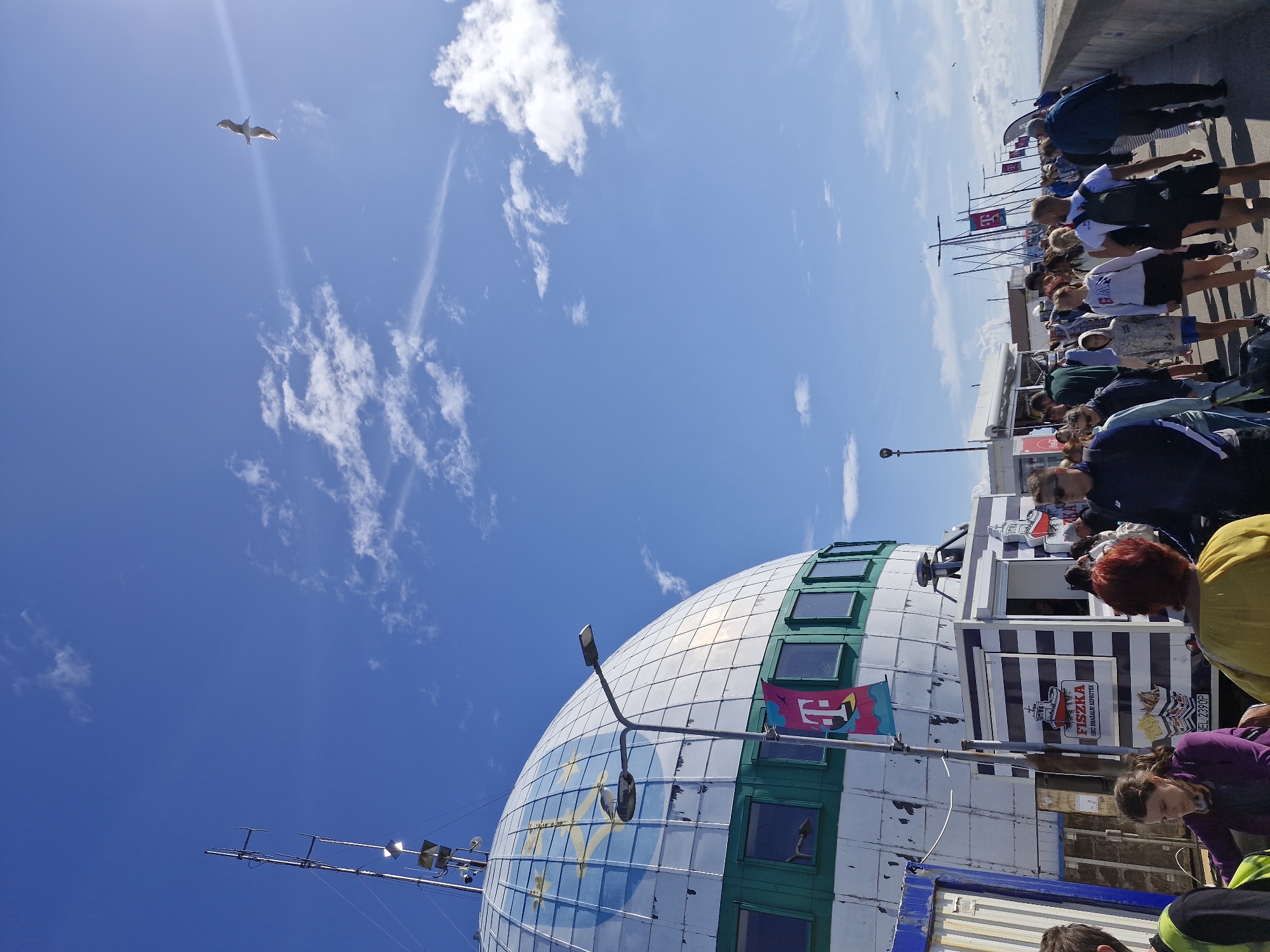 The image captures a sunny day in Hel, Poland, with a clear blue sky and scattered clouds. In the foreground, a crowd of people are walking along a paved walkway beside a large, dome-shaped building. This building appears to be a prominent landmark, possibly a cultural or historical site, with a white, segmented facade and green-trimmed windows. A T-Mobile flag is visible, suggesting a sponsorship or presence of the telecommunications company.
To the left of the dome, a food stand with a black and white striped awning is set up, featuring signage for "FISZKA" and what looks like fried fish and chips. Several individuals are queuing at the stand or being served. The people in the image are diverse in age and gender, dressed in casual summer clothing, with many carrying backpacks or bags, indicating they are likely tourists or visitors.
Further down the walkway, more people are walking, and masts of sailboats are visible in the background, suggesting proximity to a harbor or marina. The walkway itself is lined with a concrete barrier on the right, and the sea or a large body of water is visible beyond it, hinting at a coastal location. A seagull is seen flying in the sky, adding to the coastal atmosphere. The overall scene depicts a lively and bustling environment, likely a popular tourist spot in Hel.