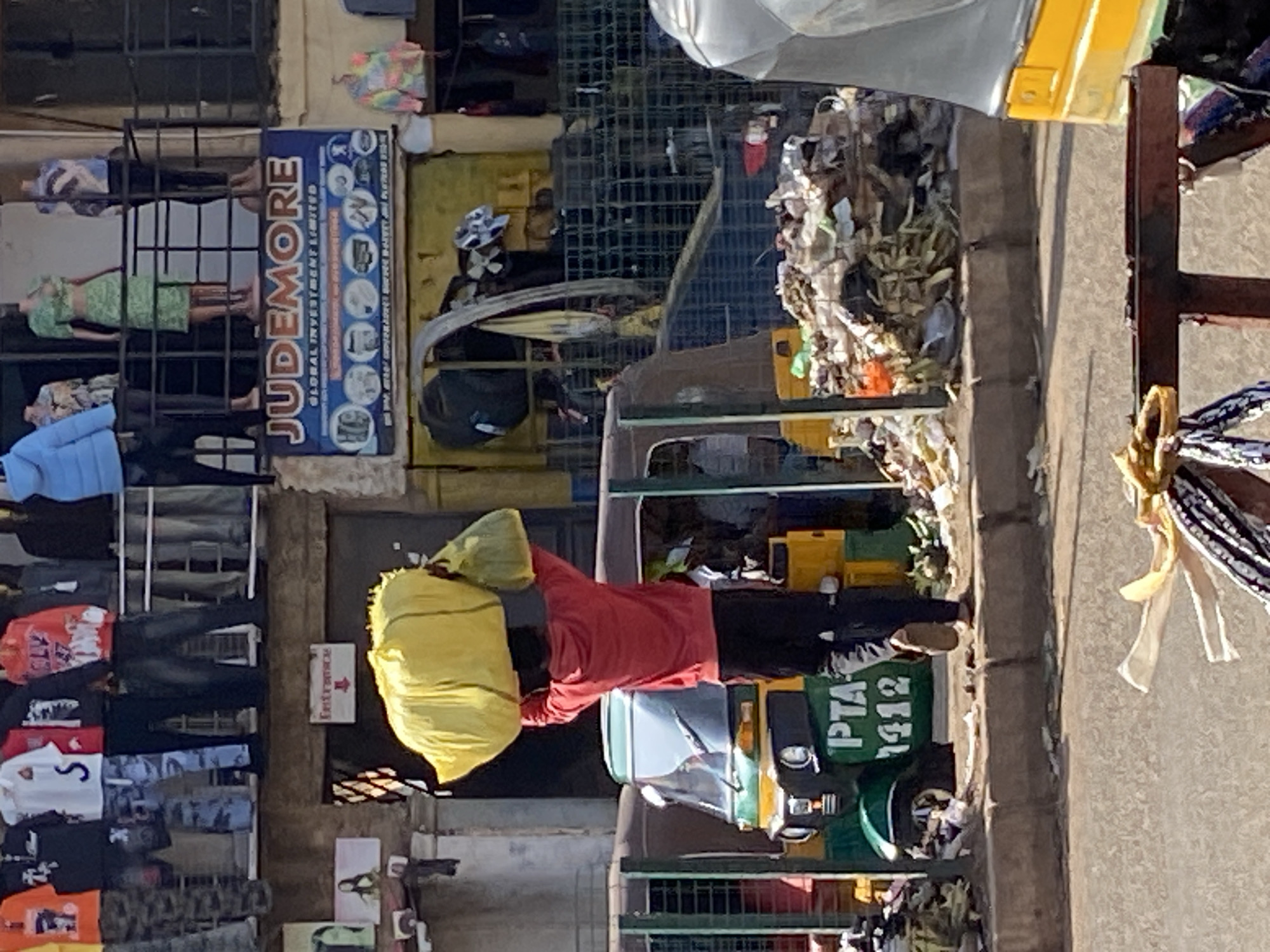 The image depicts a street scene in Bukuru, Nigeria, with a man carrying two large yellow bags on his head. He is wearing a red hooded sweatshirt and black pants. The man is standing in front of a green and yellow auto-rickshaw. The rickshaw has the text "PTA 1412" visible on its side. Behind the rickshaw is a building with a blue sign that reads "JUDEMORE GLOBAL INVESTMENT LIMITED" and various smaller circular images. A pile of trash is visible to the right of the rickshaw and building. The background shows a storefront with clothing displays and mannequins. The ground is paved and appears to be a sidewalk area.