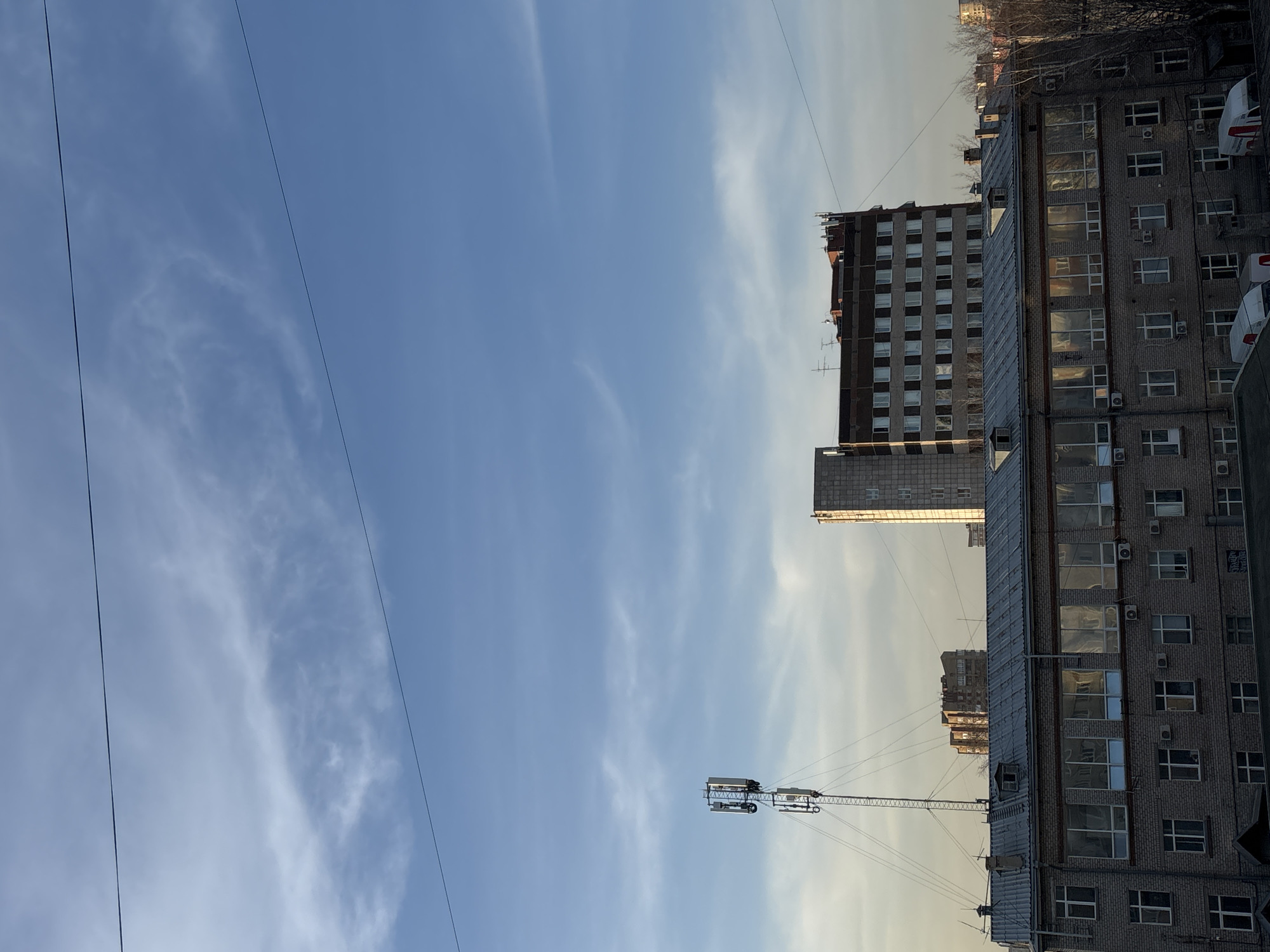 The image shows a cityscape in Perm, Russia under a clear blue sky with scattered clouds. Two utility wires run diagonally across the upper left of the frame. A gray brick building with numerous windows and air conditioning units dominates the right side of the image. Behind it, another taller gray building with a grid of windows is visible. In the foreground, a cellular tower is present. The lighting suggests it is either sunrise or sunset.