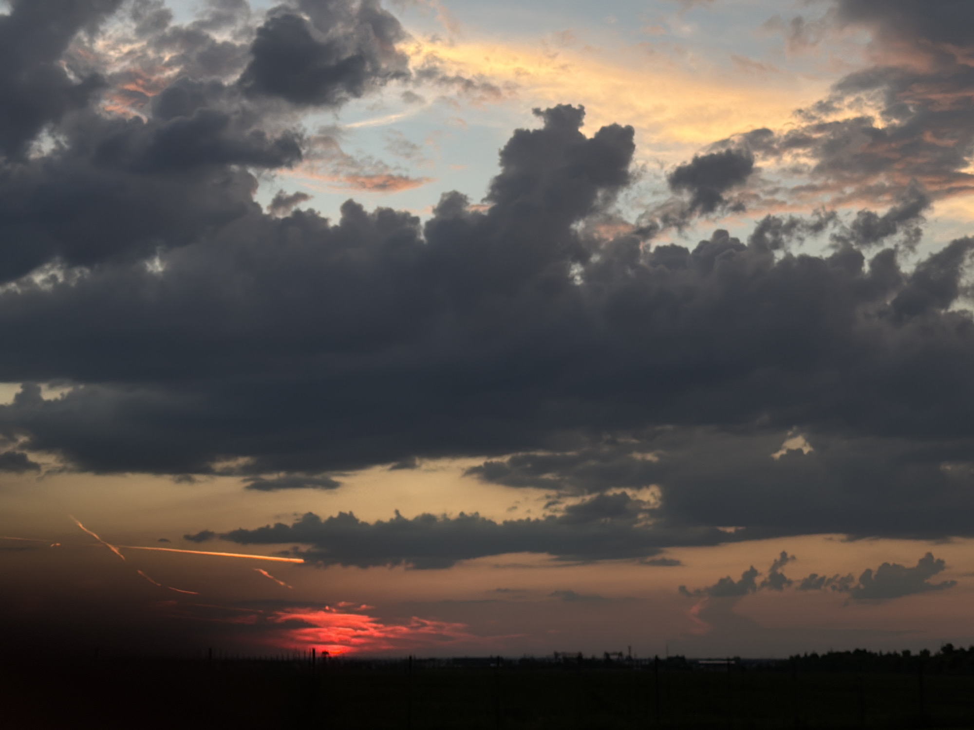 A dark, silhouetted landscape stretches across the bottom of the image, meeting a dramatic sky. The sky is filled with a thick layer of dark, heavy clouds, predominantly in shades of grey and deep blue, obscuring most of the upper atmosphere. Towards the horizon, the clouds are tinged with hues of orange and yellow, suggesting the presence of a setting sun or sunrise. A bright red glow is visible low on the horizon on the left side of the image, indicative of the sun's direct light breaking through. Streaks of orange light, possibly contrails from aircraft, cut across the sky above the red glow. The overall scene is one of a cloudy, atmospheric dusk or dawn over a rural or open area, possibly fields or plains, with a dark silhouette of vegetation or a very low tree line along the horizon. The location is identified as Timofeyevka, Russia, implying a vast, open landscape typical of the region under this dramatic sky.