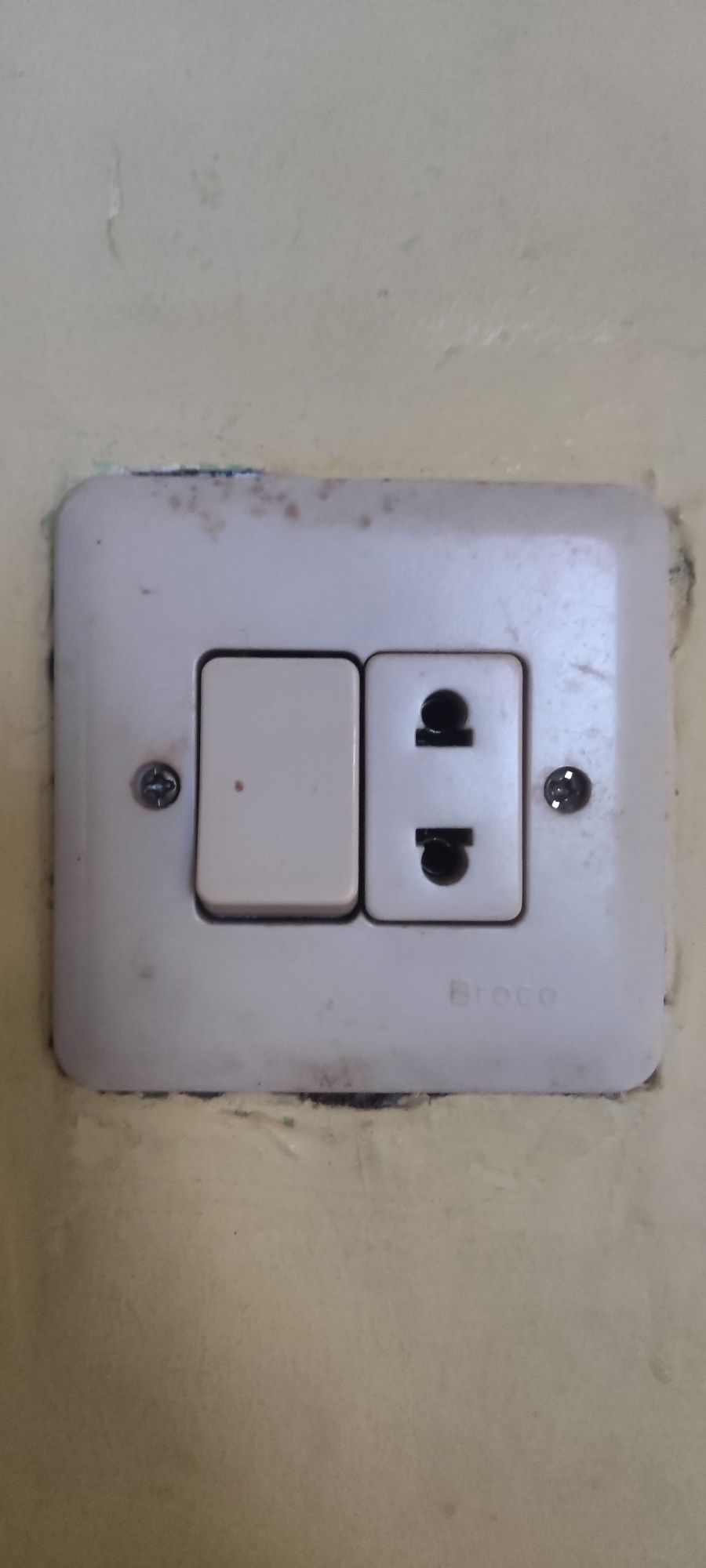 The image displays a close-up view of a white electrical outlet and switch mounted on a light yellow wall. The outlet is a universal type, capable of accepting various plug configurations, and features two circular openings with a ground pin slot in between. To the left of the outlet is a rectangular light beige switch. The entire unit is secured to the wall by two visible screws, one on the left side and one on the right side of the plate. The surface of the outlet and switch plate shows some signs of age and wear, with smudges and a few small dark specks, suggesting it may not be new. The wall itself appears to have a textured finish and exhibits some staining and discoloration, particularly around the edges of the electrical fixture. The brand name "Broco" is faintly visible embossed on the bottom right of the outlet plate. There are no people or actions depicted in this image. The scene appears to be an interior electrical fitting, likely in a residential or commercial building. Based on the prompt's request to include the city of Gebang, Indonesia, this electrical fitting is located there.
